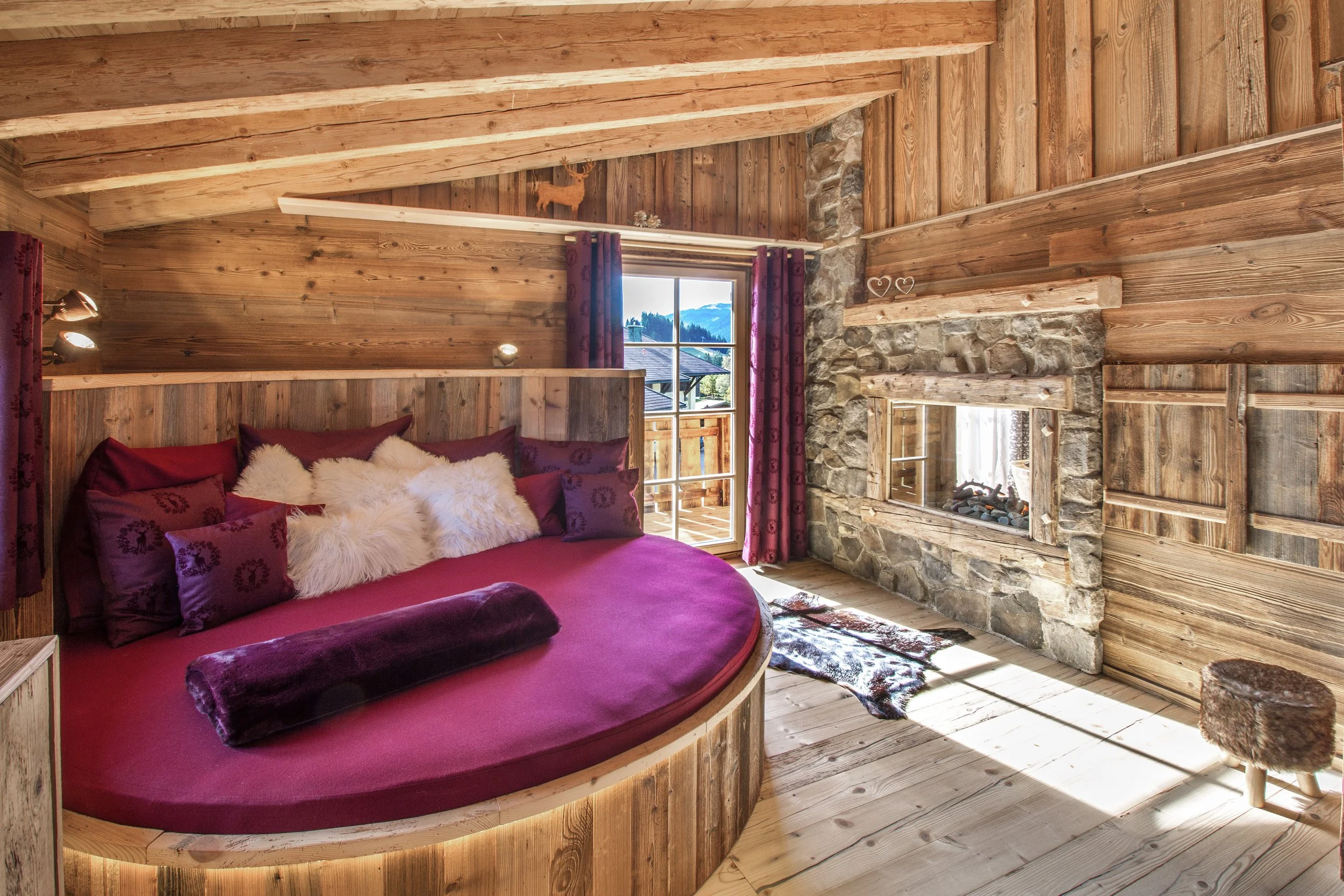 Gemütliches Schlafzimmer mit Holzvertäfelung, rotem Bett, gefilzten Kissen, Fenster mit Blick auf Berge, Kamin aus Stein, Herzdekorationen auf der Mantel, Vorhänge in Dunkelrot, Holzfußboden.