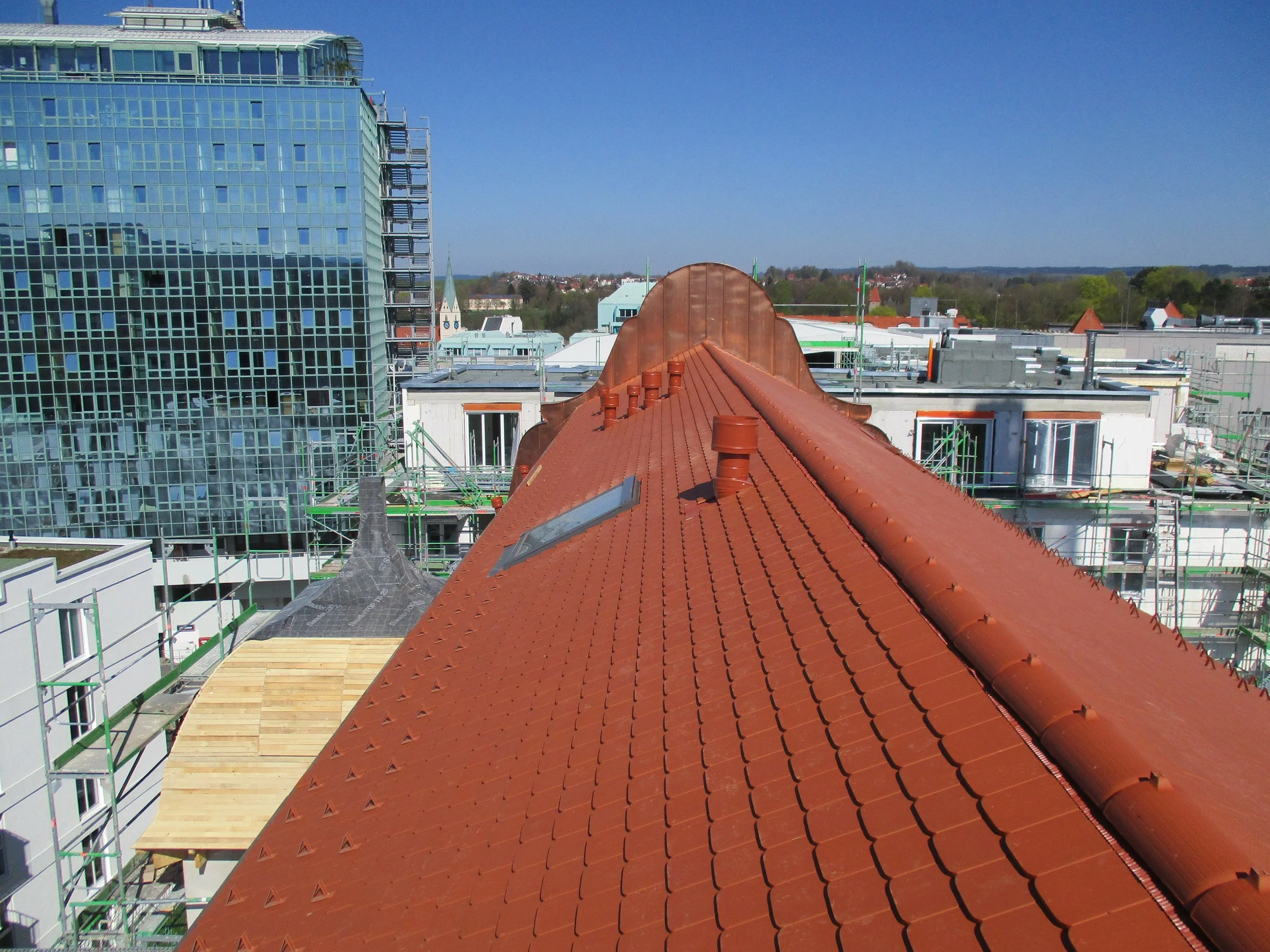 Blick auf eine Baustelle auf einem Stadtdach mit rot-weißen Ziegeln, Baugerüsten und einem modernen Glasgebäude im Hintergrund, sowie einem blauen Himmel.