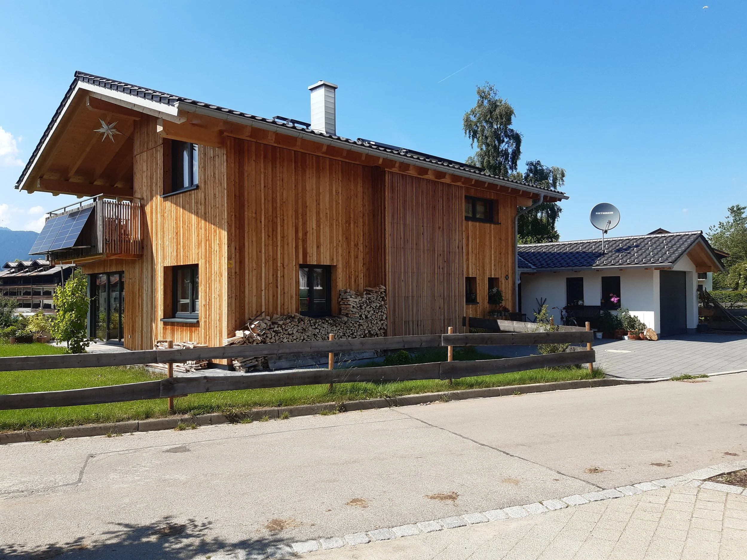 Ein mehrstöckiges Holzhaus mit Dach, einer Metallziegeldeckung, einem Kamin, einem Balkon, Solarzellen und einem Satellitenantenne. Im Hintergrund sind Bäume und ein blauer Himmel sichtbar.