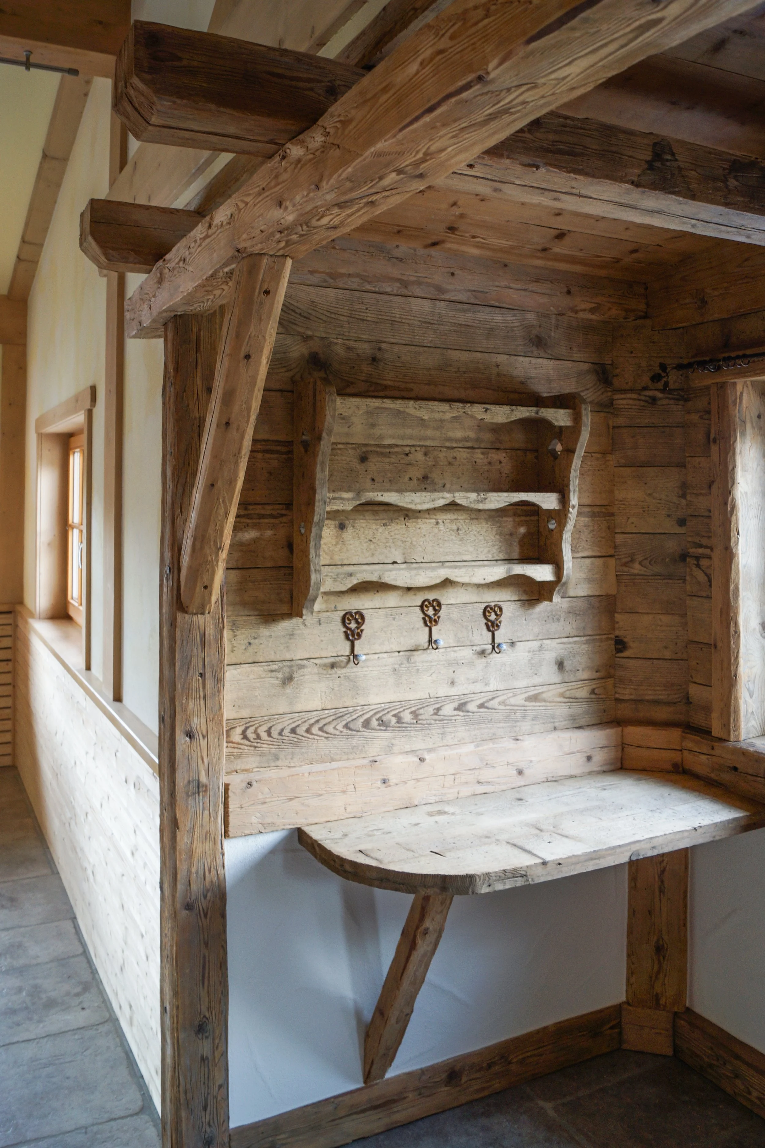Rustikaler Holzwand mit Regal, Haken und kleiner Holzbank, in einem Raum mit Steinboden und Fenster.