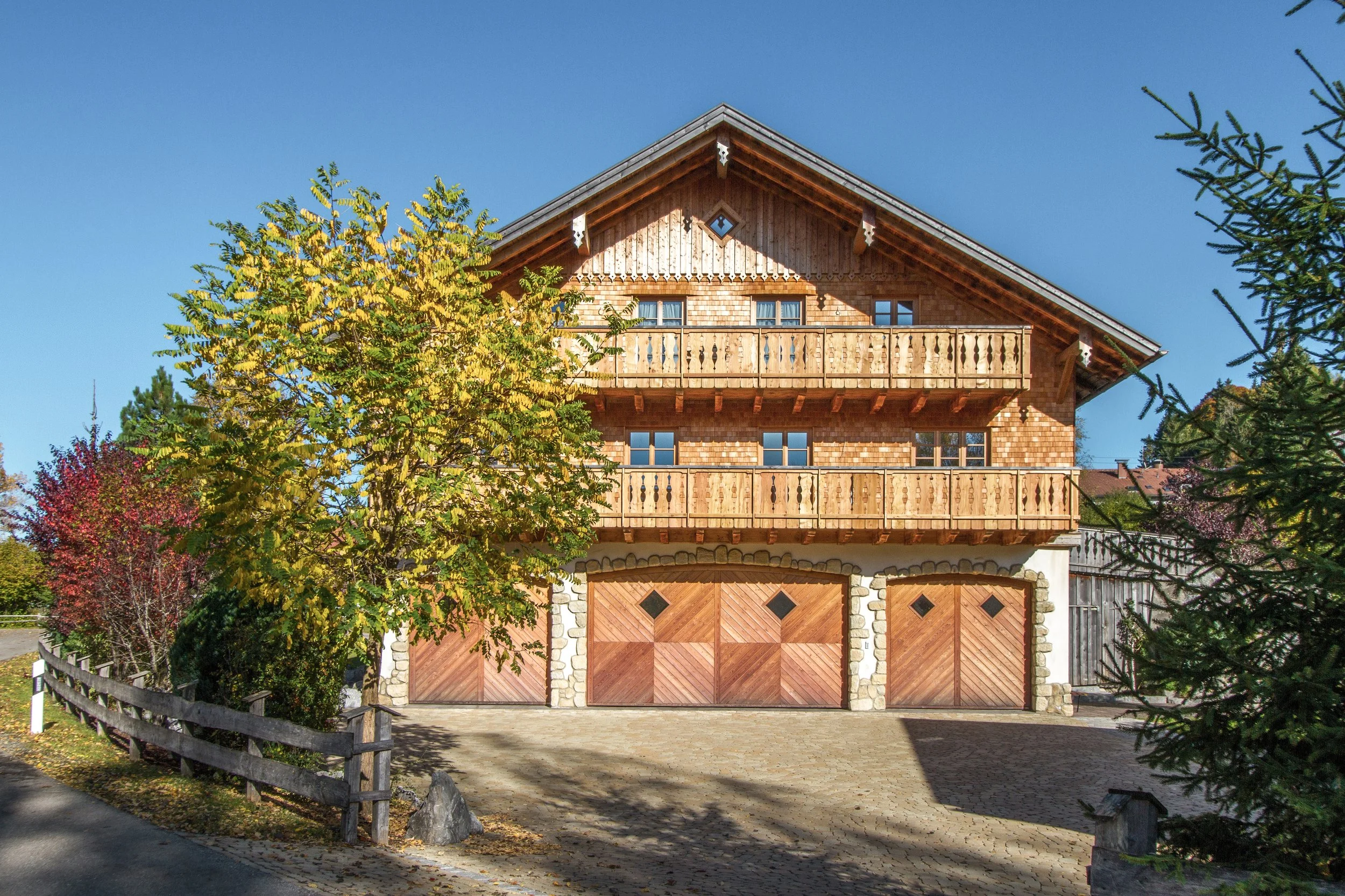 Ein Haus mit großen hölzernen Balkonen, umgeben von Bäumen mit gelben und grünen Blättern, bei sonnigem Wetter im Herbst.