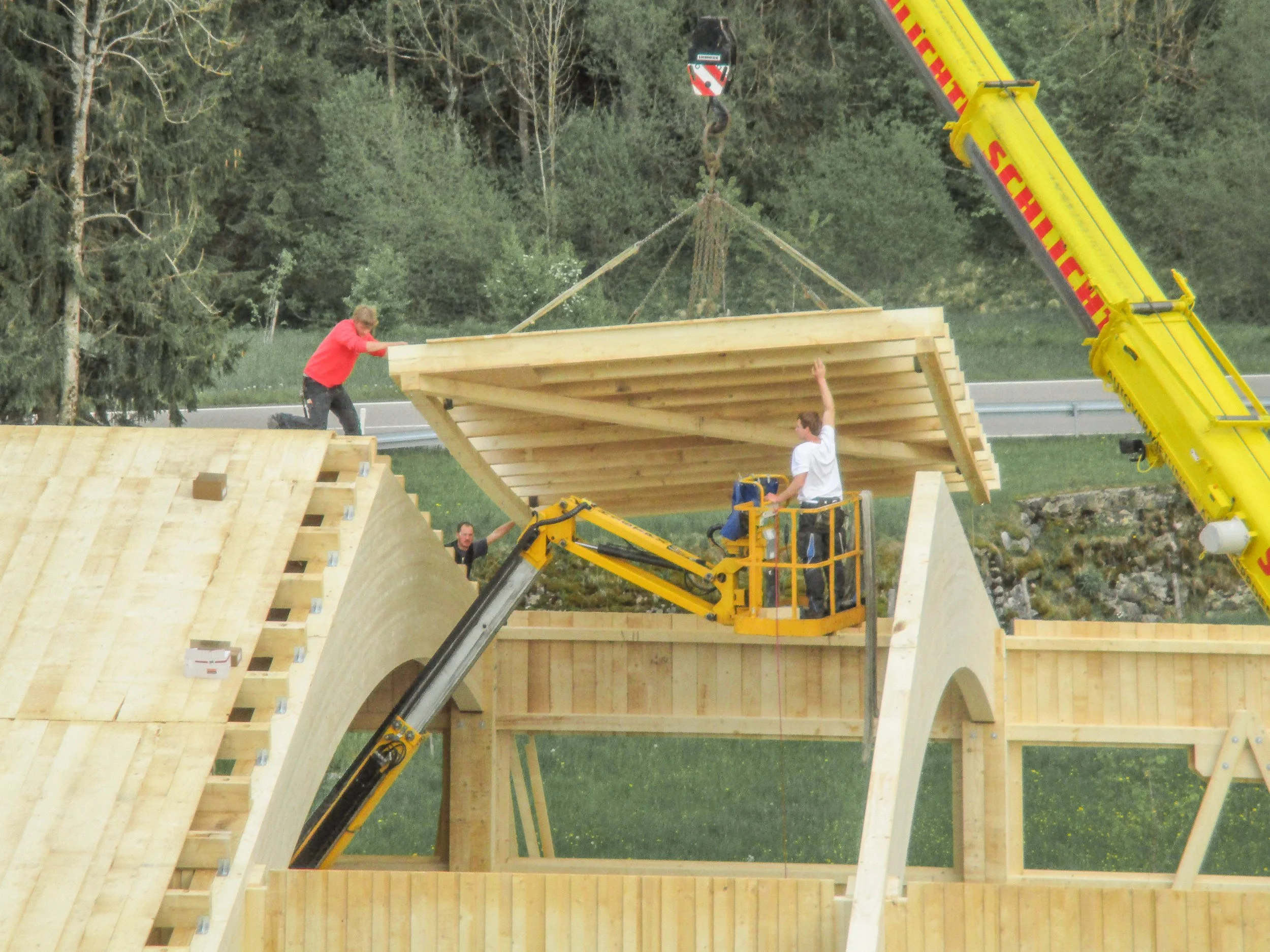 Menschen bauen Holzrahmen für ein Gebäude mit einer Kranmaschine und einer Baustellenbühne. Es sind Bäume im Hintergrund sichtbar.