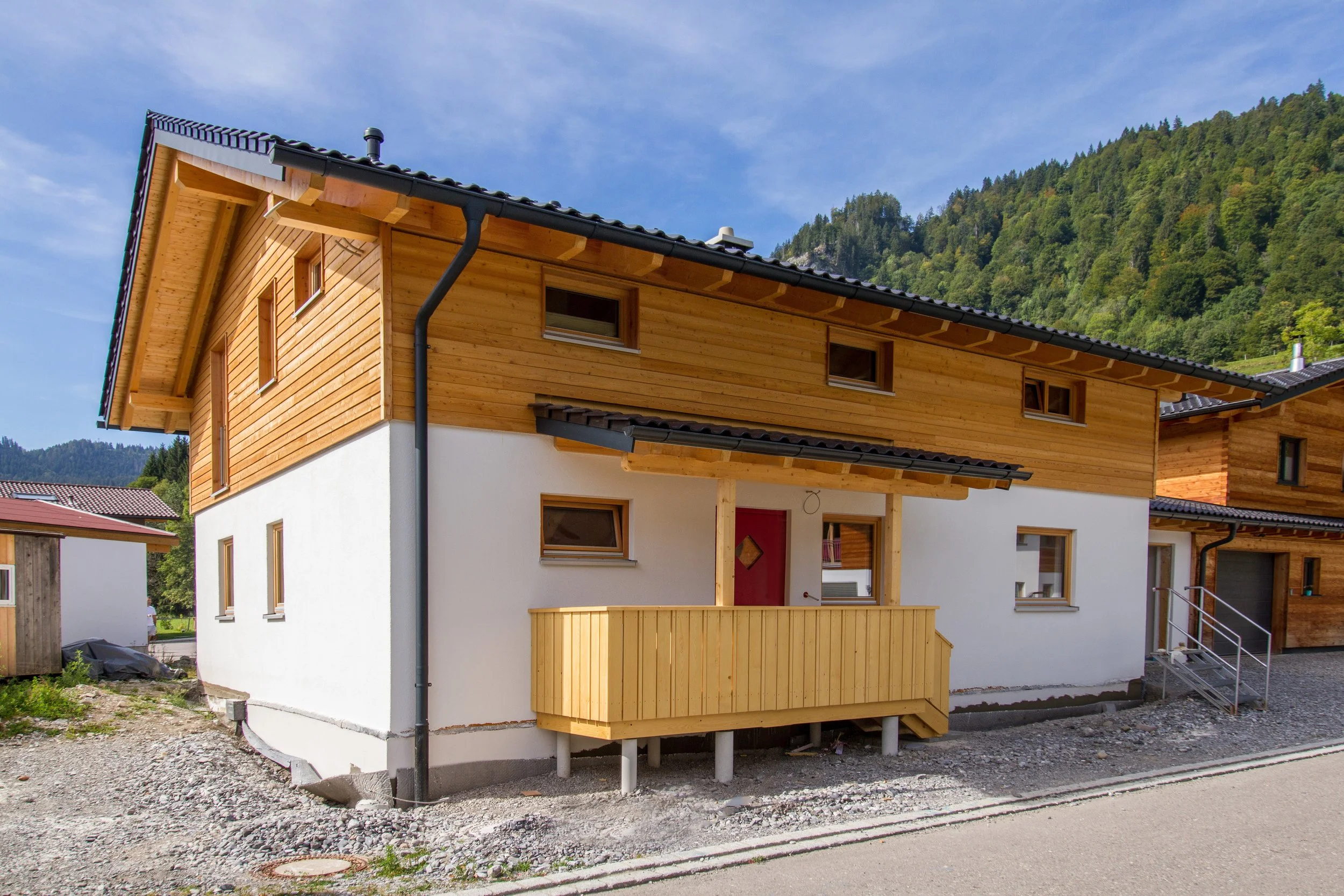 Neues zweistöckiges Haus mit Holzfassade und weißen unteren Wänden, gebaut vor einem bewaldeten Berg im Hintergrund, bei schönem Himmel.