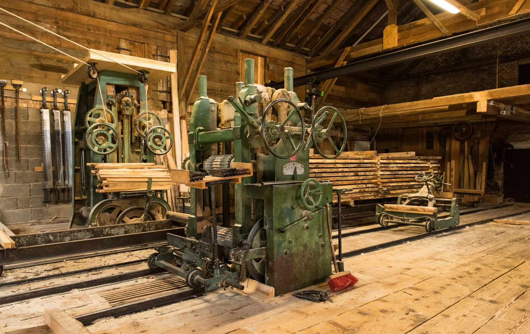 Ein Holzbearbeitungsraum mit einer alten, grünen Holzspaltmaschine, mehreren Holzstapeln und verschiedenen Werkzeugen an der Wand.