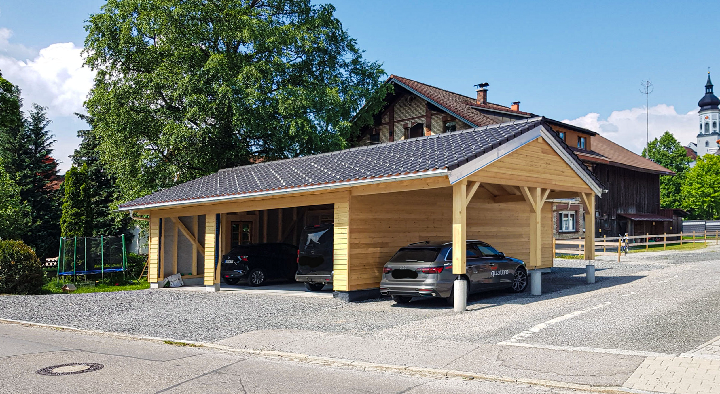 Ein Carport mit Holzrahmen und Ziegeldach, darunter Parken ein schwarzer Audi, ein grauer Opel und ein schwarzes Fahrzeug. Im Hintergrund sind Bäume, ein Haus und eine Kirche sichtbar.