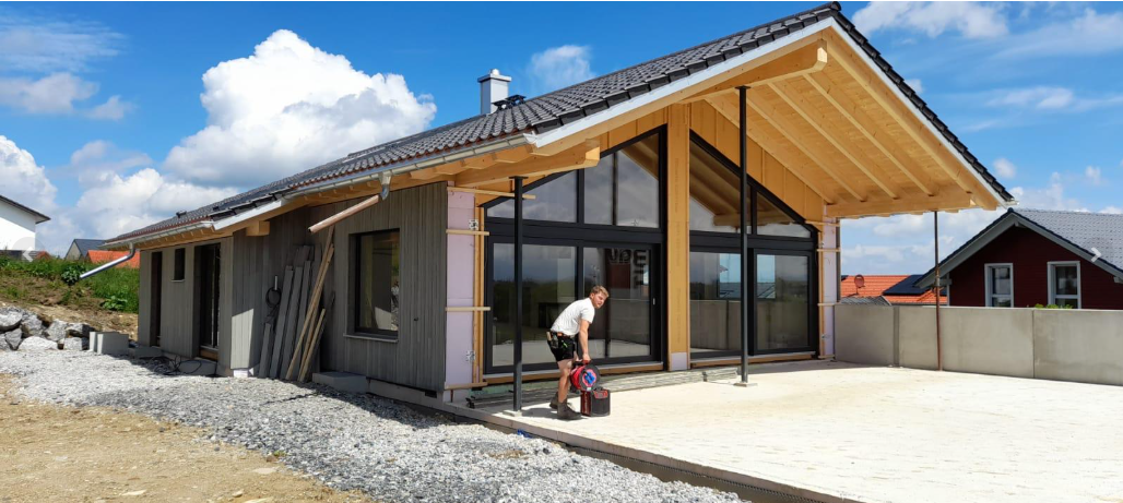 Ein Haus im Bau mit holzverkleideter Fassade, große Glasfenster und einem großen überdachten Balkon, im Hintergrund sind Wolken am Himmel sichtbar.