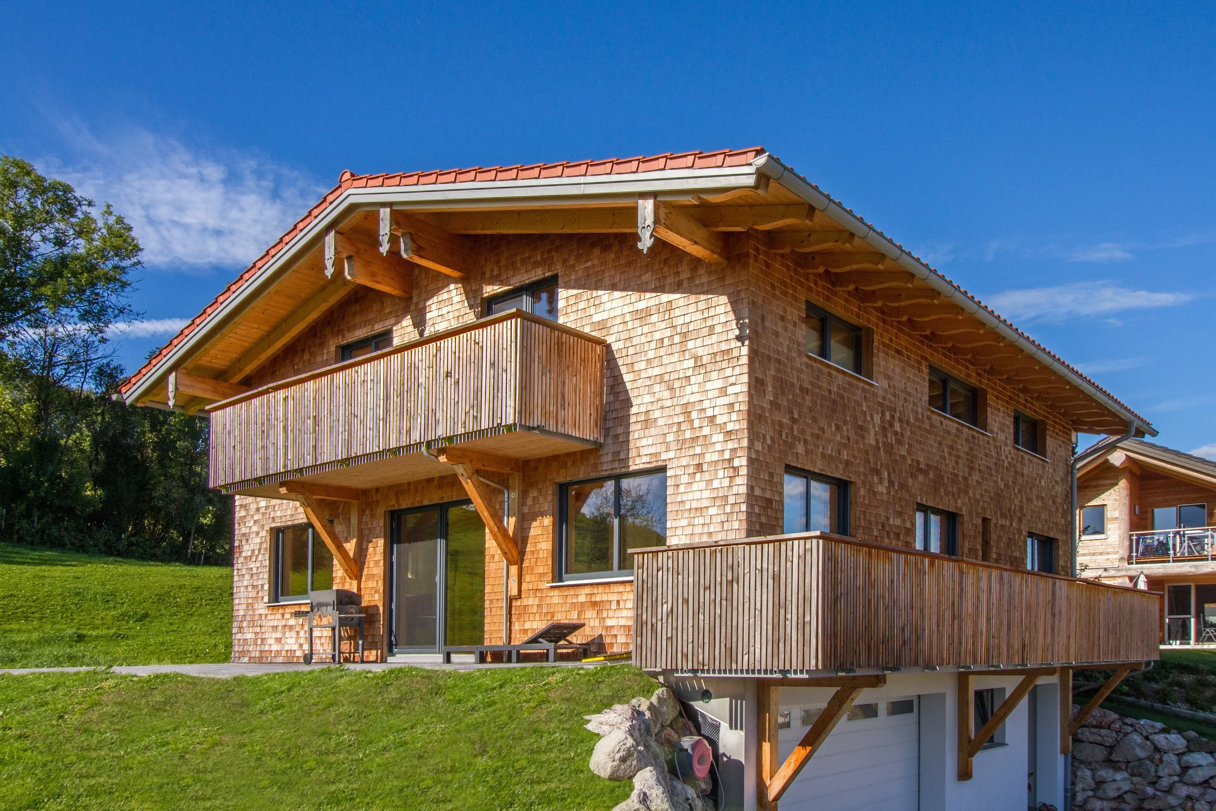 Mehrstöckiges Haus mit Holzfassade und roten Dachziegeln, Balkon, große Fenster, grüne Rasenfläche, blauer Himmel, umgeben von Bäumen.