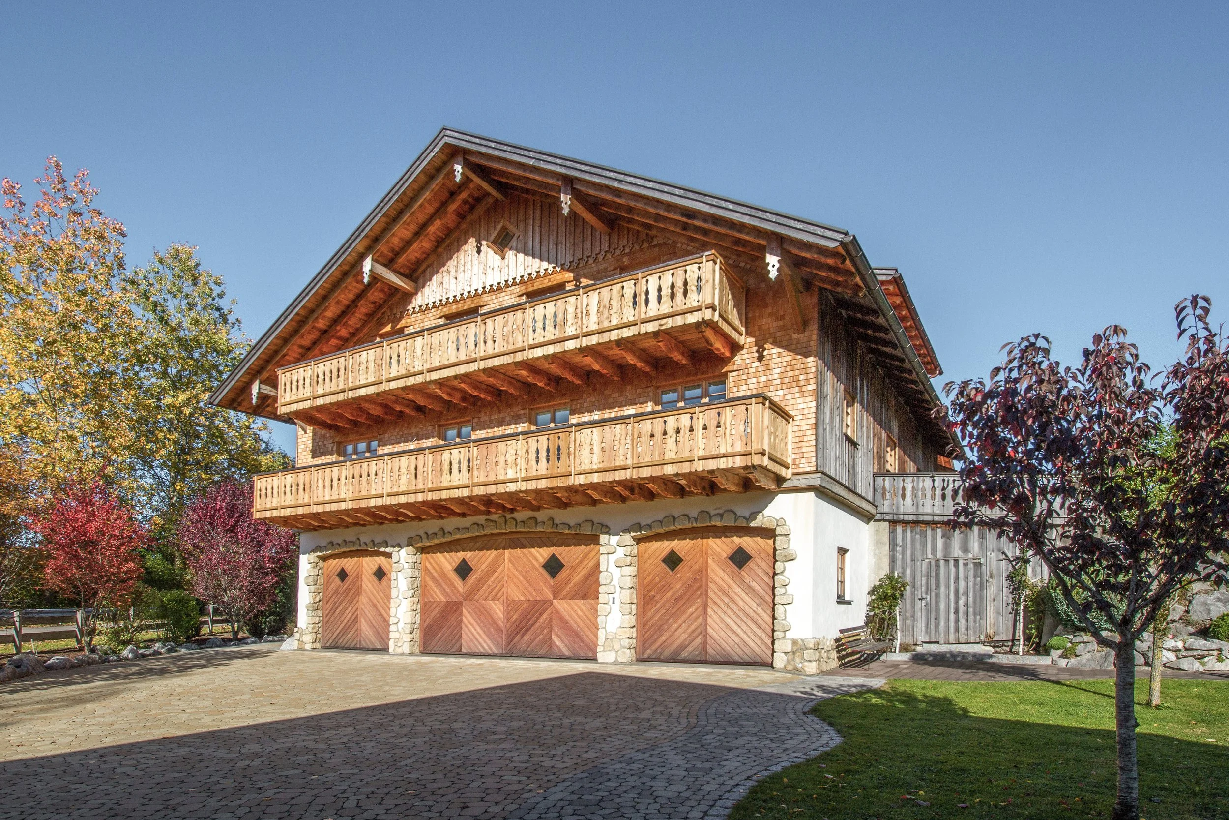 Holzhaus mit drei Garagentoren und Balkon, umgeben von Bäumen mit Herbstlaub, im klaren blauen Himmel.