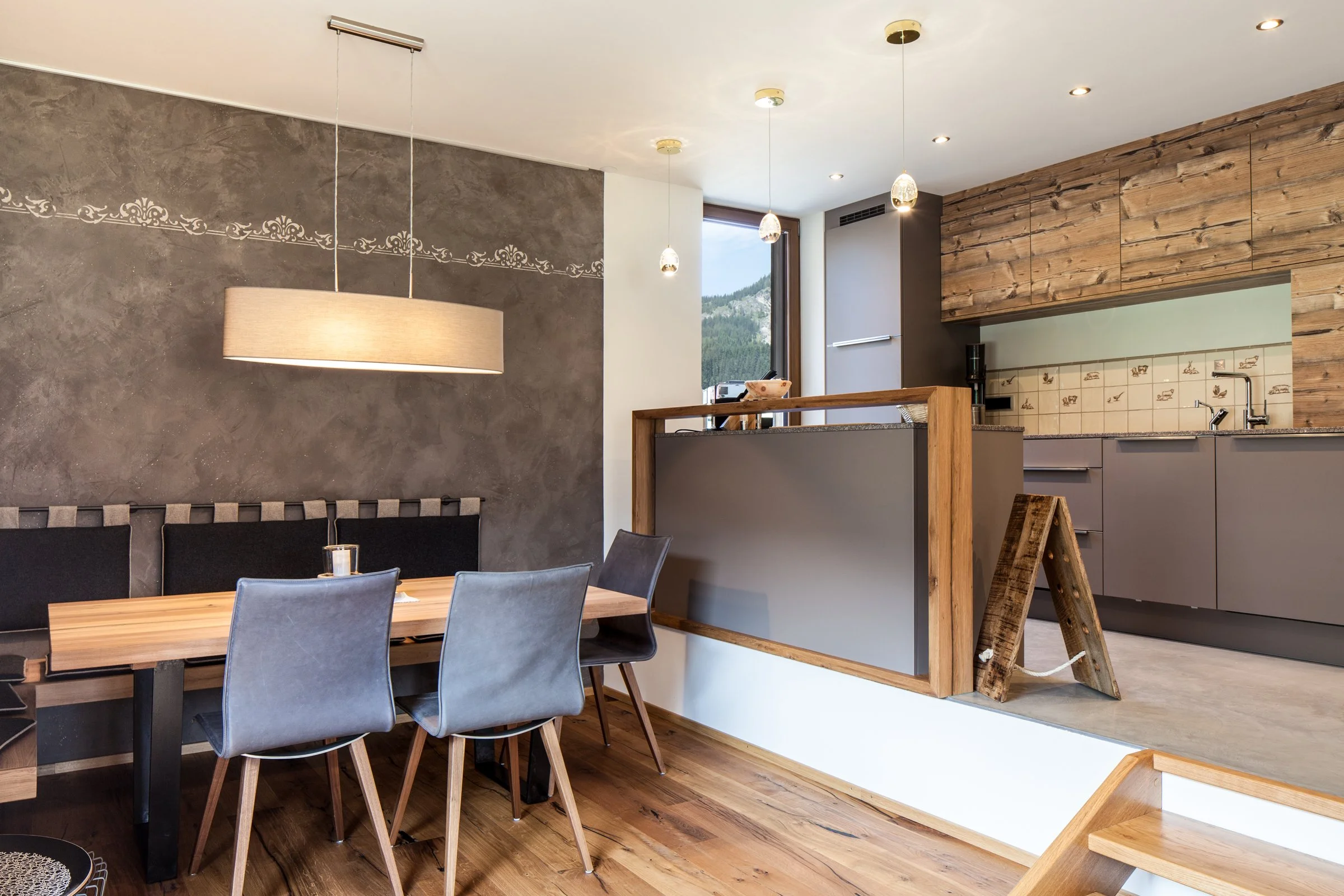 Ein moderner Essbereich mit einem Holztisch, mehreren grauen Stühlen, einer grauen Wand mit beschriebener Verzierung und einer Holzwand im Küchenbereich im Hintergrund, mit Blick auf eine große Fensterfront und Berge im Außen.