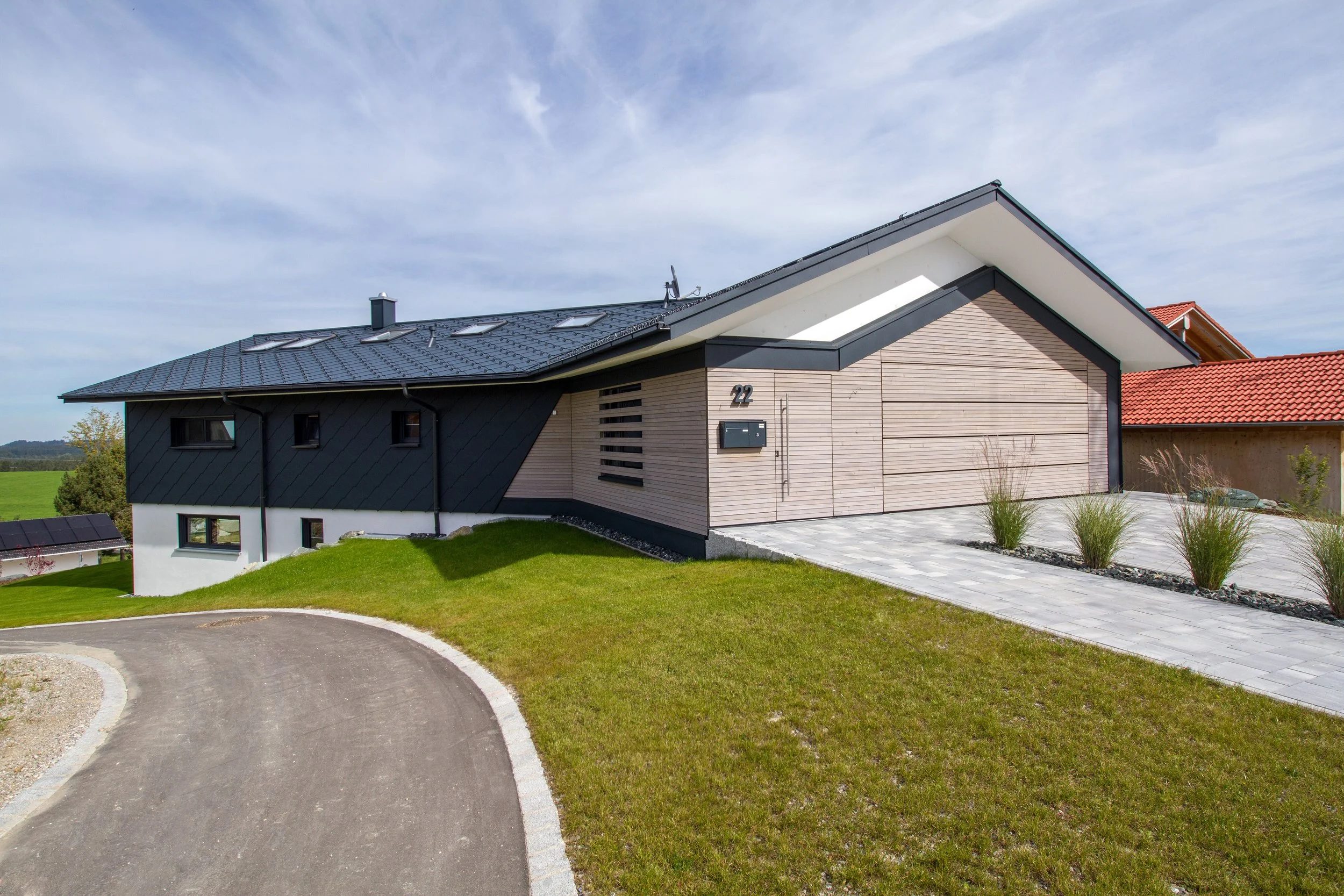 Modernes Haus mit schwarzem und hellem Holzfassade, welligem Dach mit Solarpanelen, vor einer grünen Wiese und blauem Himmel.