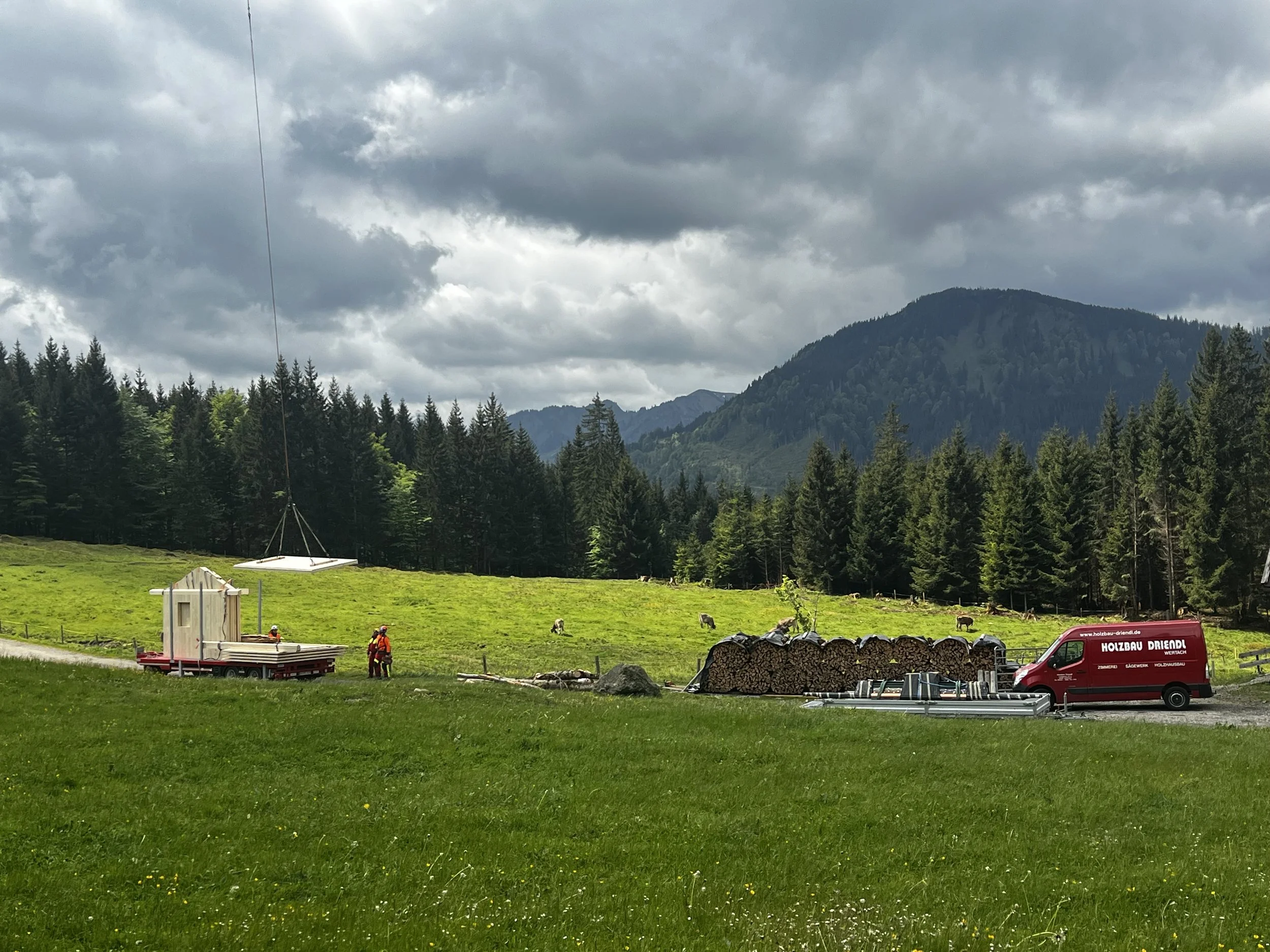 Grünes Wiesengelände im Vordergrund mit einigen Helfern, Holzstapeln und Fahrzeugen, umgeben von dichten Wäldern und Bergen bei bewölktem Himmel.