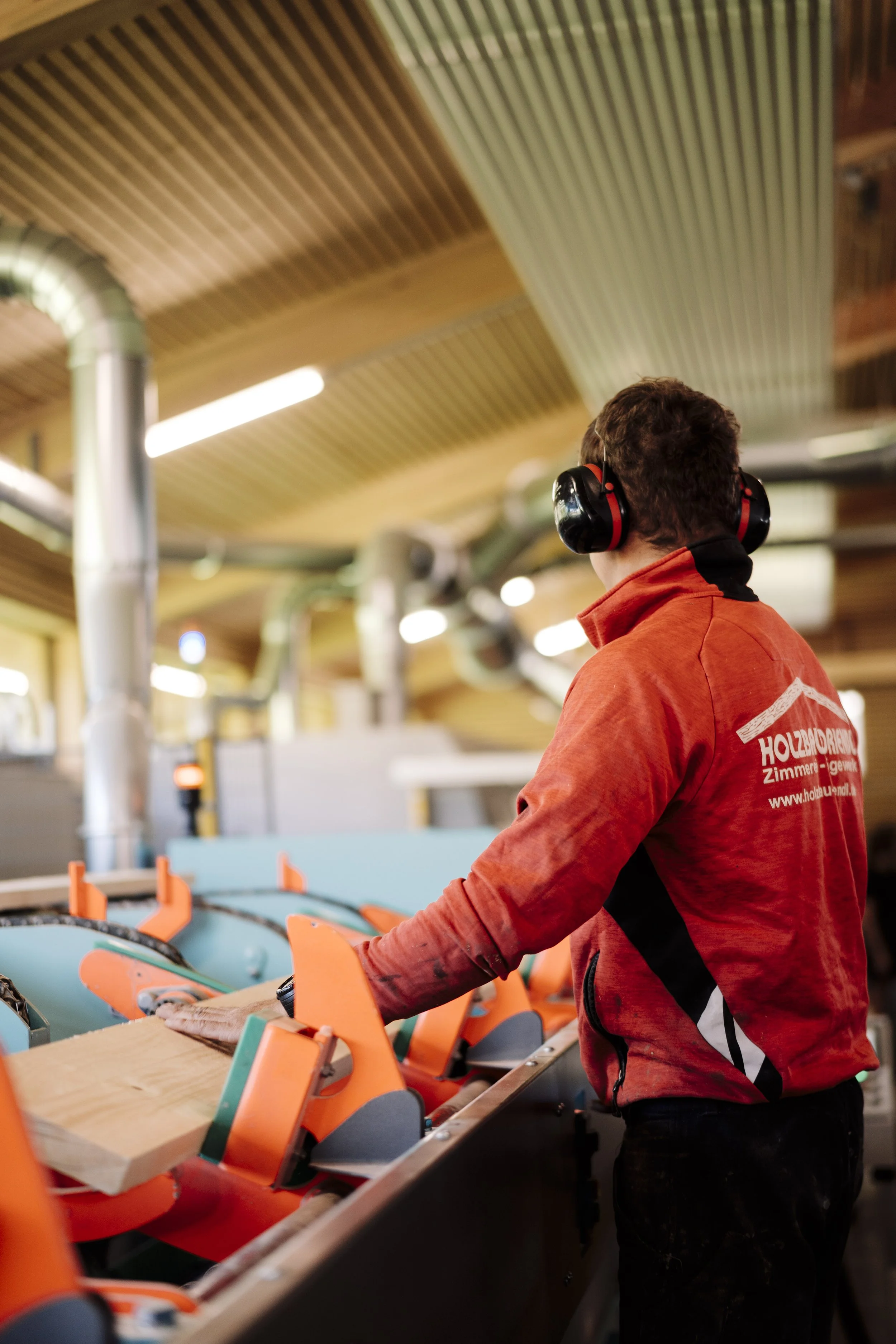 Ein Mann trägt ein rotes Jacke mit Druck auf der Rückseite und hört mit schwarzen Gehörschutz, während er eine Maschine in einer Holzwerkstatt bedient.