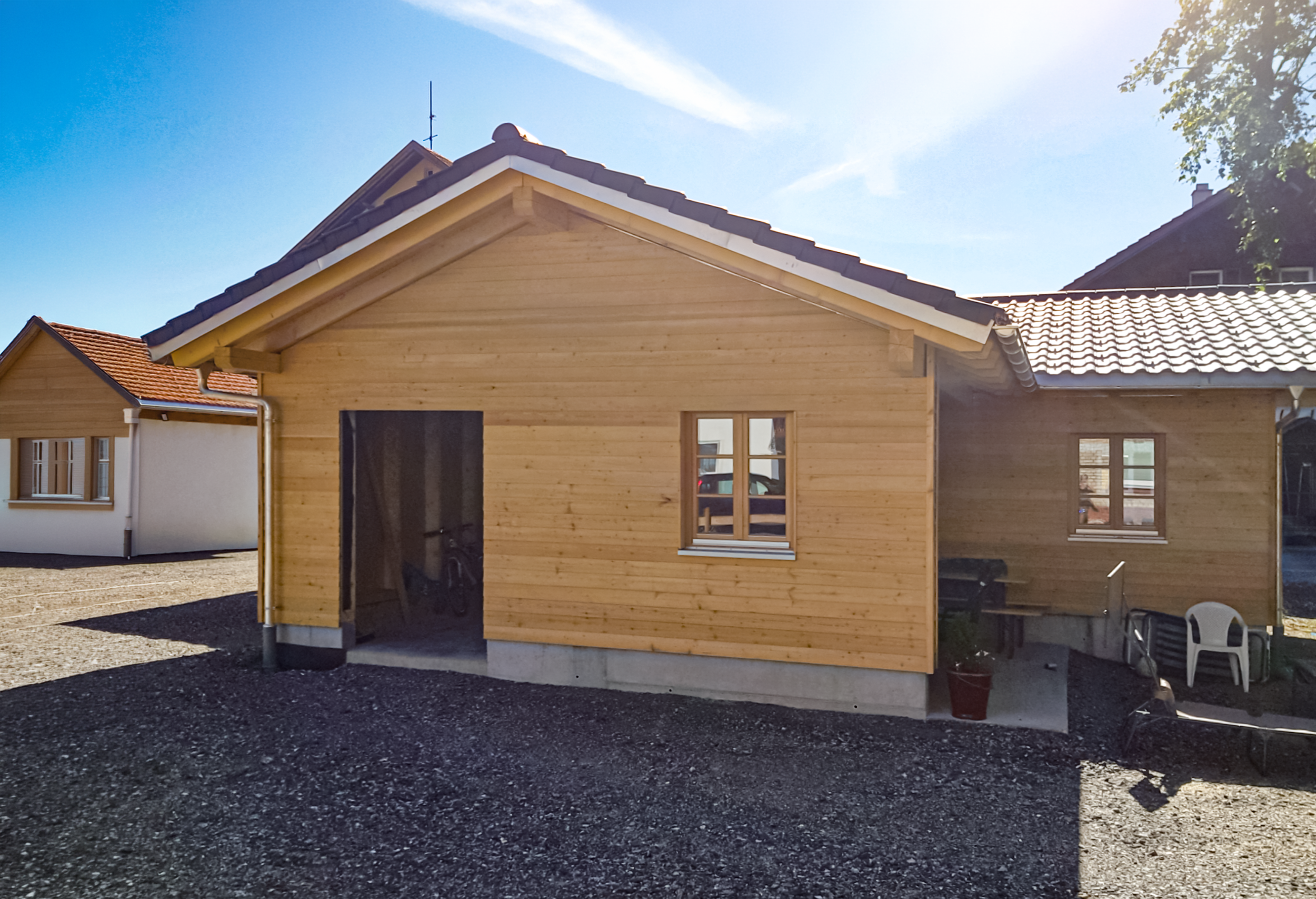 Ein neu gebautes Holzhaus auf einem unbefestigten, schwarzen Kiesgrund bei Sonnenlicht, mit einem blauen Himmel im Hintergrund.