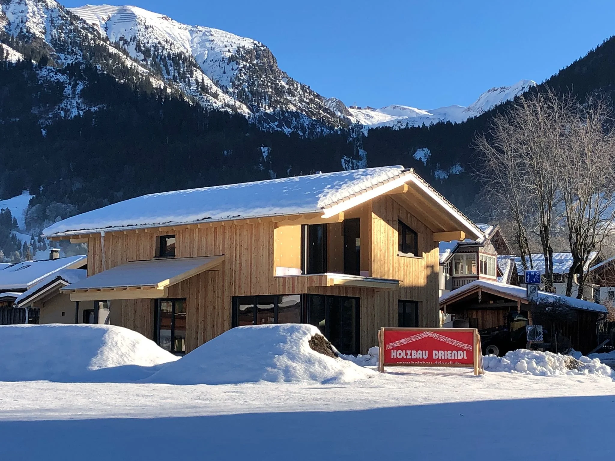 Ein modernes Holzhaus mit großen Fenstern in einer verschneiten Berglandschaft, umgeben von schneebedeckten Bäumen und Bergen unter klarem blauen Himmel.