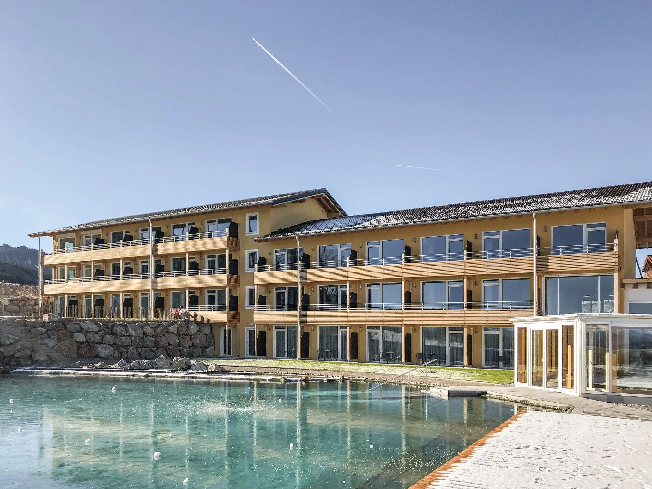 Modernes Hotelgebäude mit mehreren Balkonen, Blick auf den Hotelpool im Vordergrund, Wasser, und einen klaren blauen Himmel mit Flugzeugspoiler im Hintergrund.