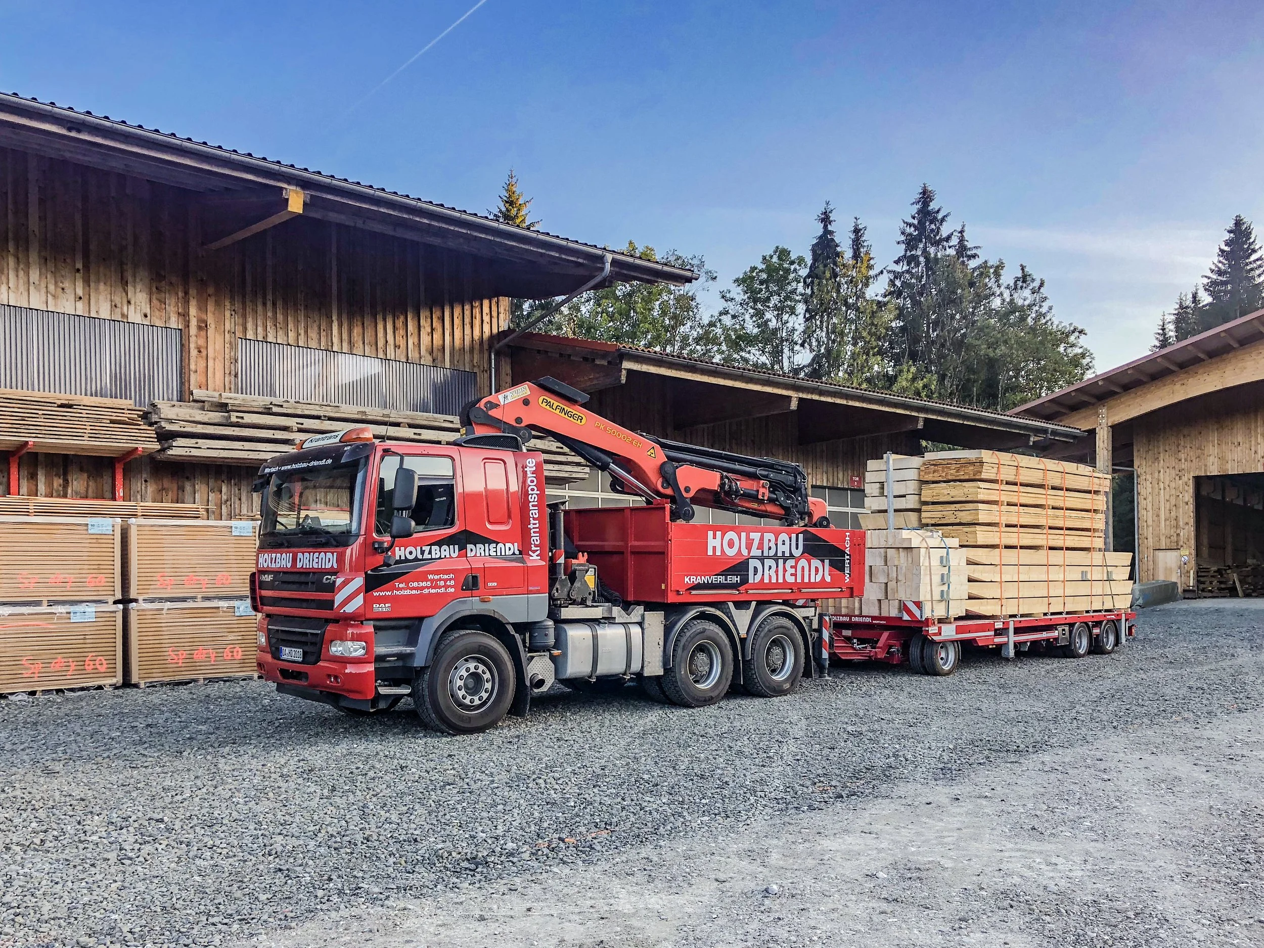 Ein rotes Baukranfahrzeug auf Kiesboden, das Baumaterialien wie Holzplatten und Balken transportiert, vor einem Holzbauernhof mit Bäumen im Hintergrund.