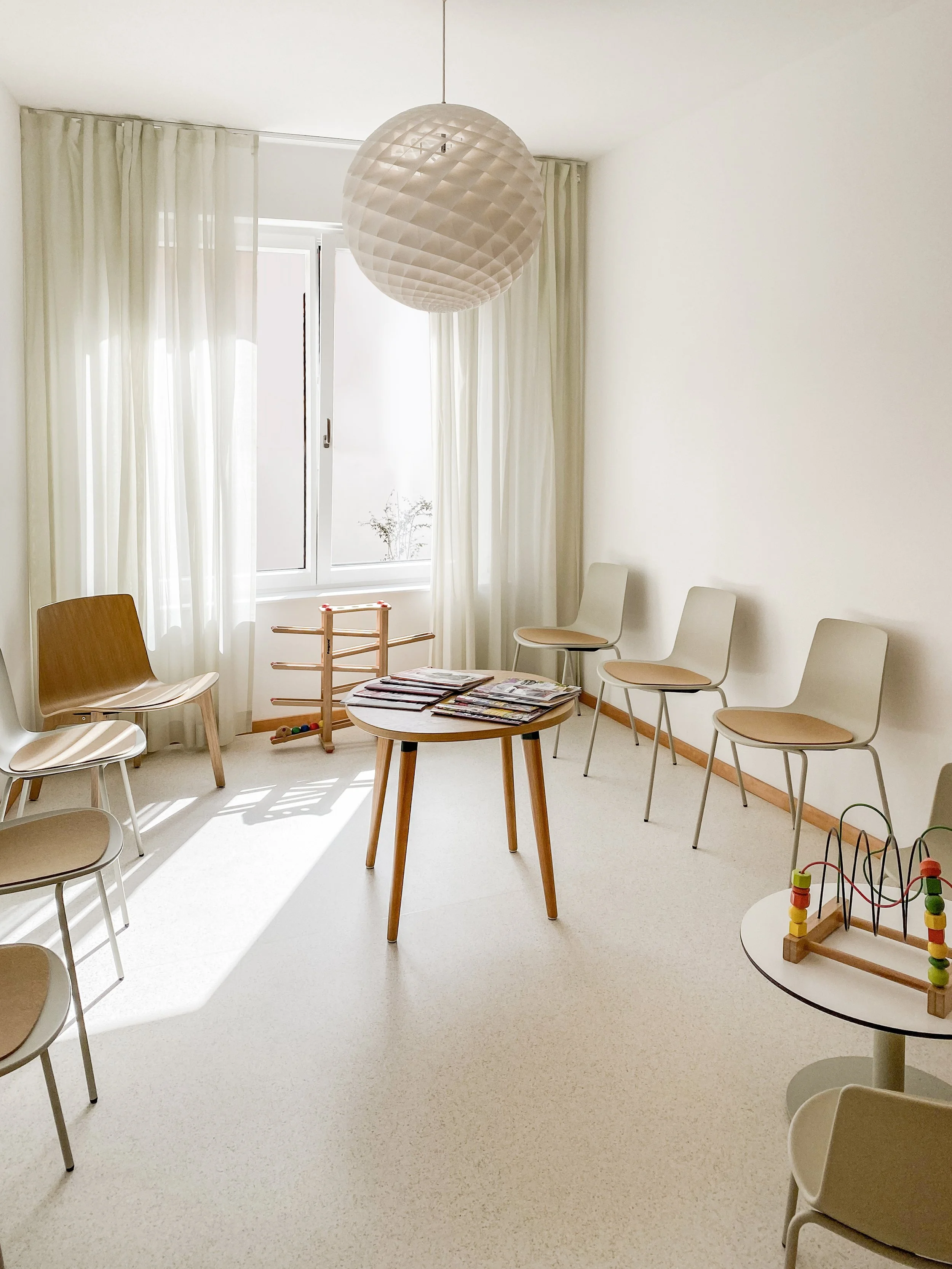 Ein heller Raum mit weißen Wänden, mehreren Stühlen, einem Tisch mit Magazinen, einem Hängependel-Licht, einem Fenster mit weißen Vorhängen und einigen Spielzeugen auf einem kleinen Tisch.