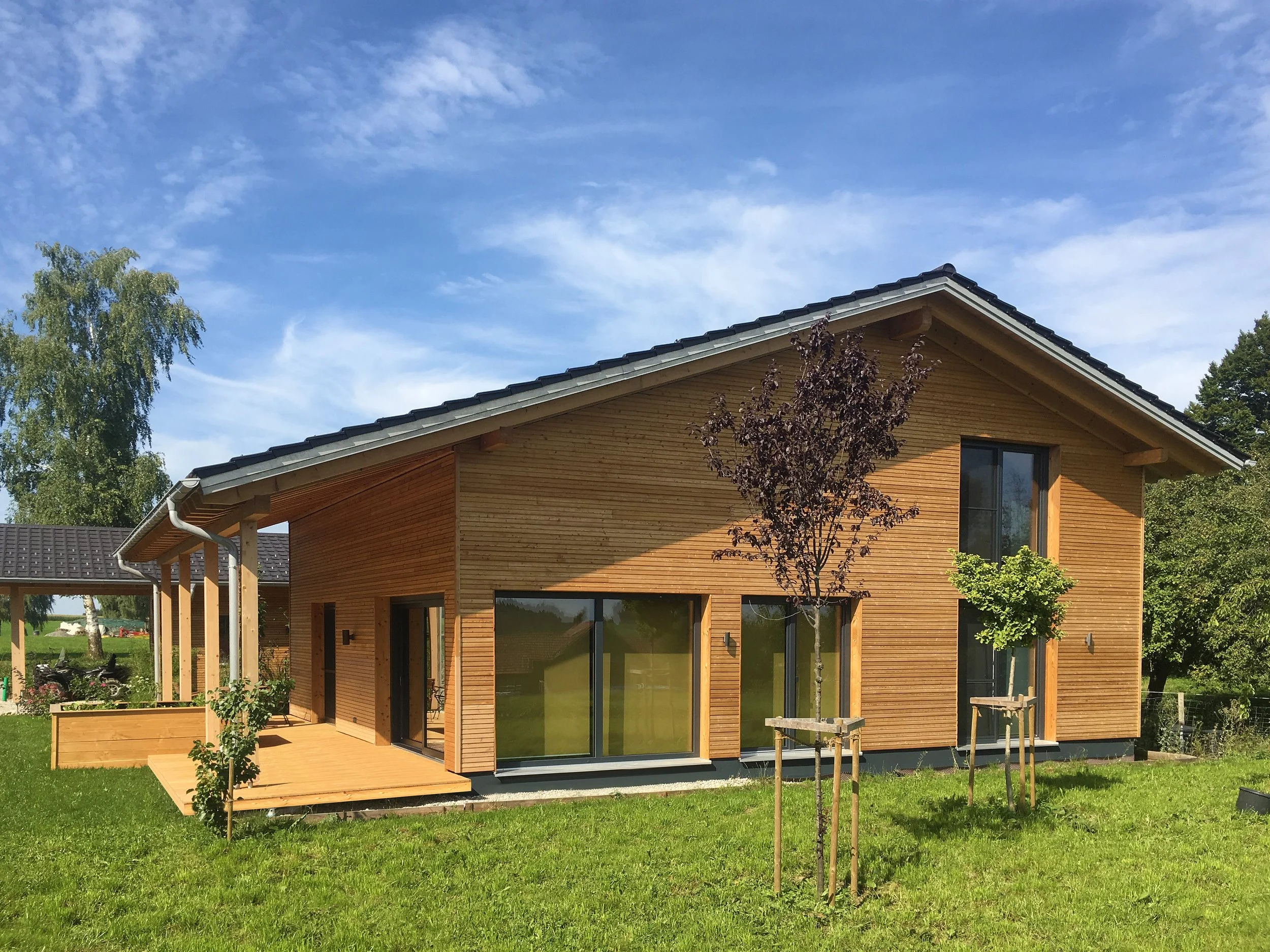 Ein modernes Holzhaus mit großen Fenstern und einem Balkon im Garten, umgeben von Bäumen und Grünsland, bei blauem Himmel mit wenigen Wolken.