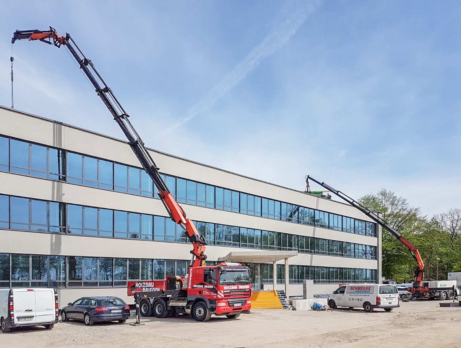 Bauarbeiten an einem modernen vierstöckigen Bürogebäude mit großen Glasscheiben, bei denen Kräne auf dem Dach installiert werden.