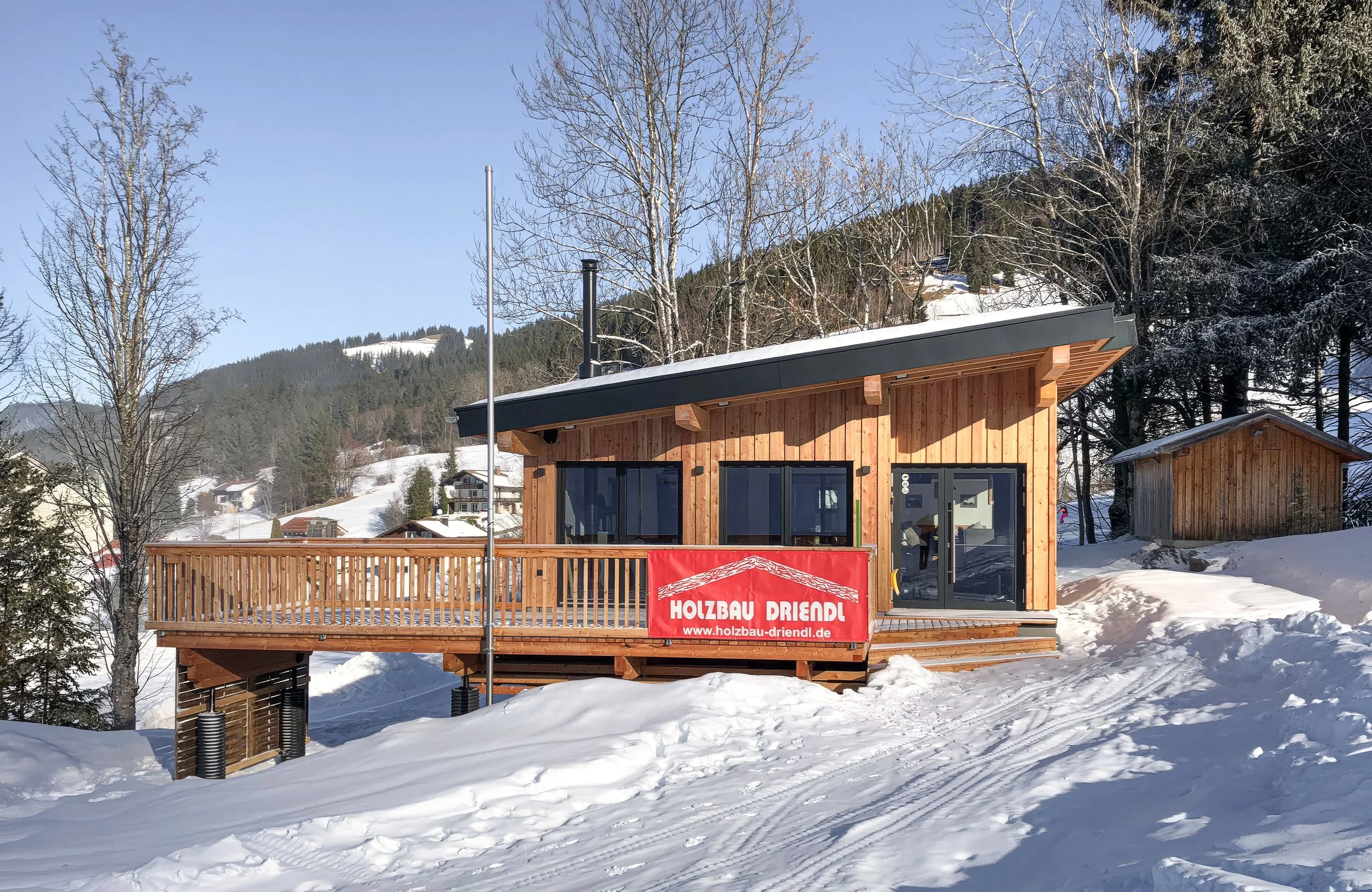 Holzhaus mit Terrasse in verschneiter Winter Landschaft, umgeben von Bäumen und Bergen, mit Werbung für Holzbau Driendl
