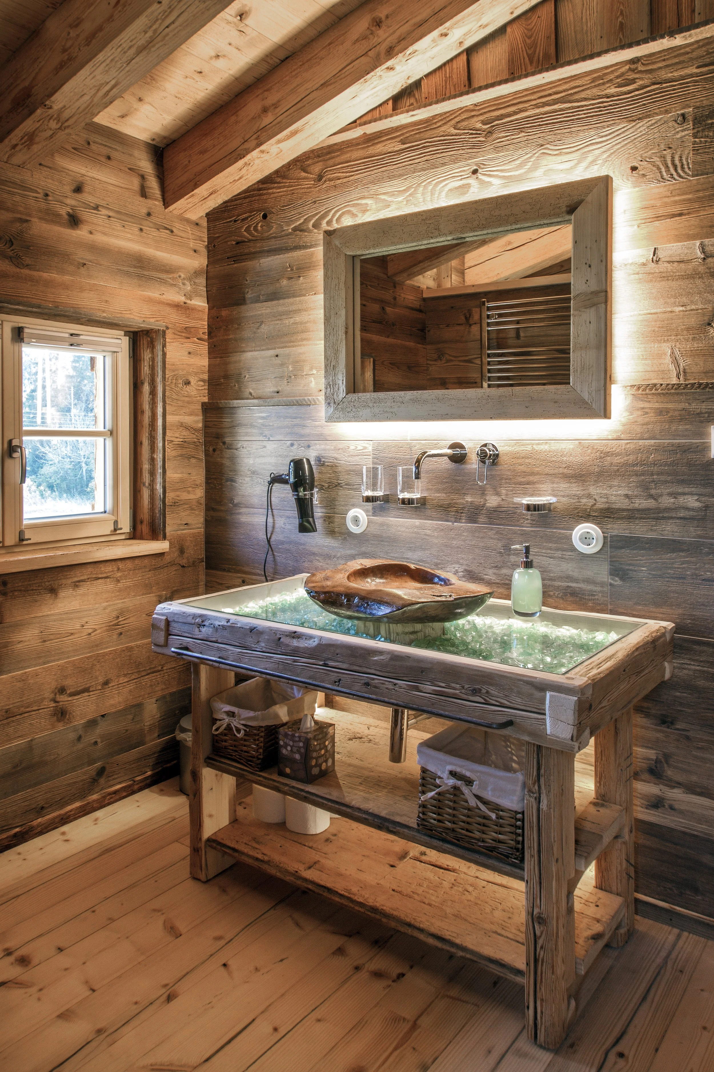 Ein Badezimmer mit Holzverkleidung, Waschbecken aus natürlichem Holz, einer großen Spiegelwand mit Beleuchtung, einem kleinen Fenster und einem Handtuchheizkörper.