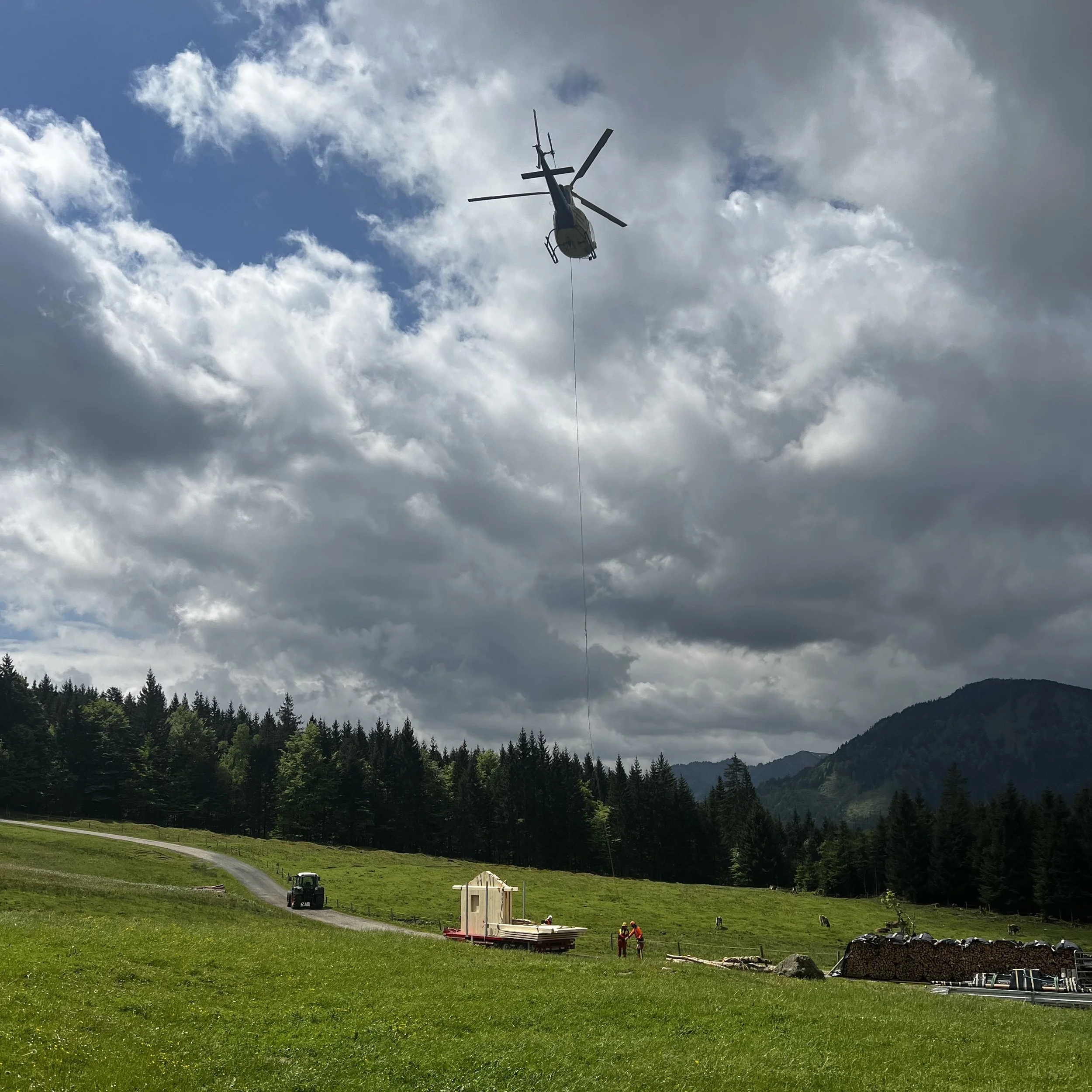 Blick auf eine grüne Wiese mit Wald im Hintergrund, ein Traktor, Baumstämme, Arbeitsgeräte, und Personen die bei Bauarbeiten sind. Im Himmel eine Hubschrauber mit sichtbarer Seilwinde, Wolken und Berge im Hintergrund.