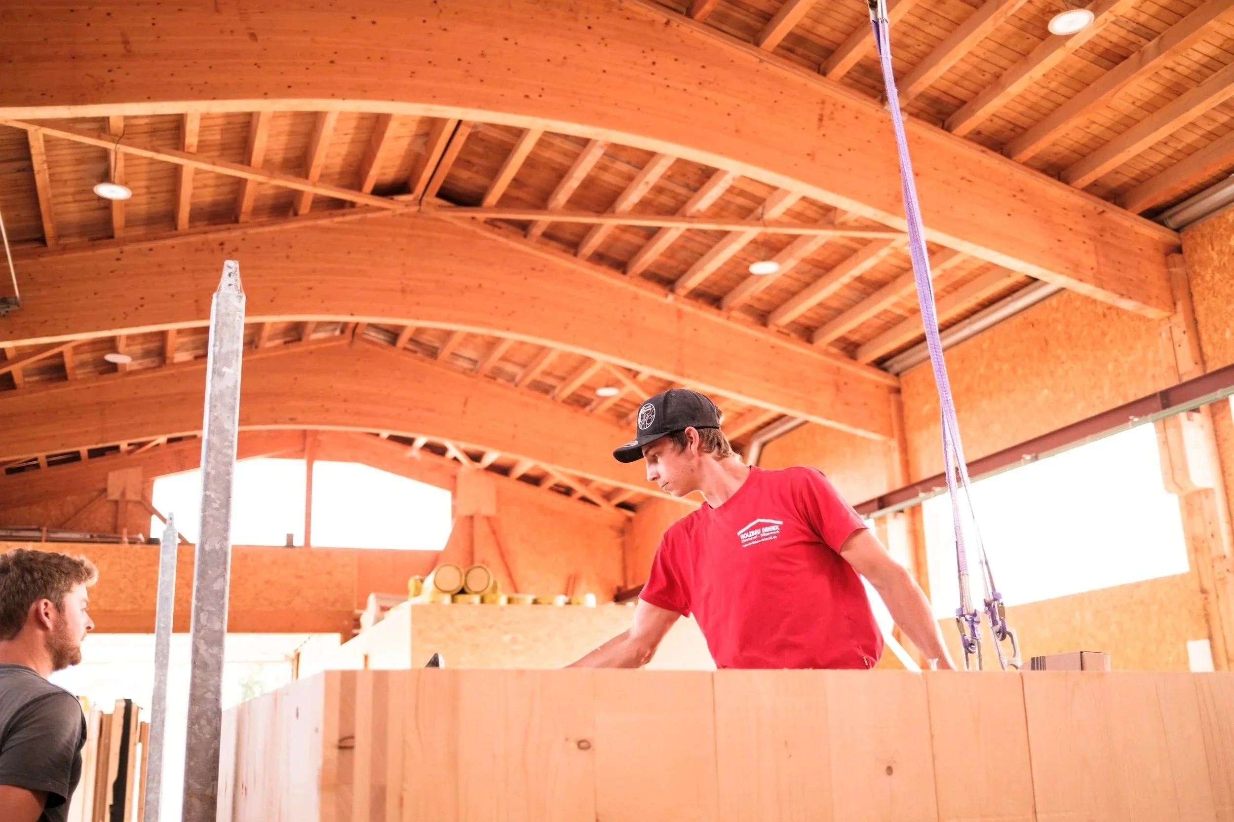 Ein Holzhaus mit gewölbter Holzbalkendecke im Inneren, zwei Männer, einer trägt eine schwarze Mütze und ein rotes T-Shirt, der andere sitzt mit braunen Haaren. Es scheint eine Baustelle oder Werkstatt zu sein.