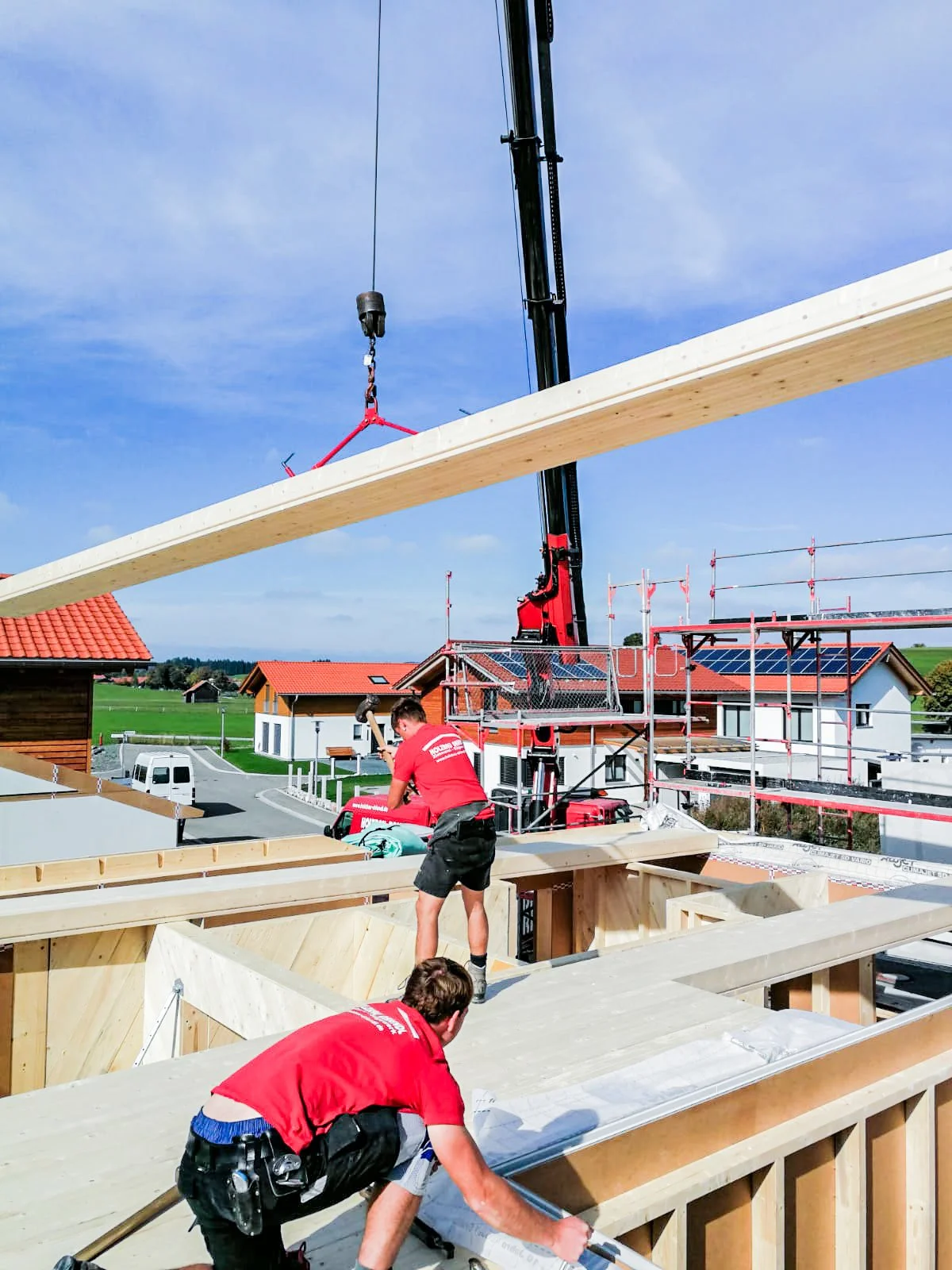 Menschen bauen Holzrahmen für ein Haus, während ein Kran ein Holzstück hebt. Das Wetter ist sonnig und es befinden sich moderne Häuser mit Solaranlagen in der Umgebung.