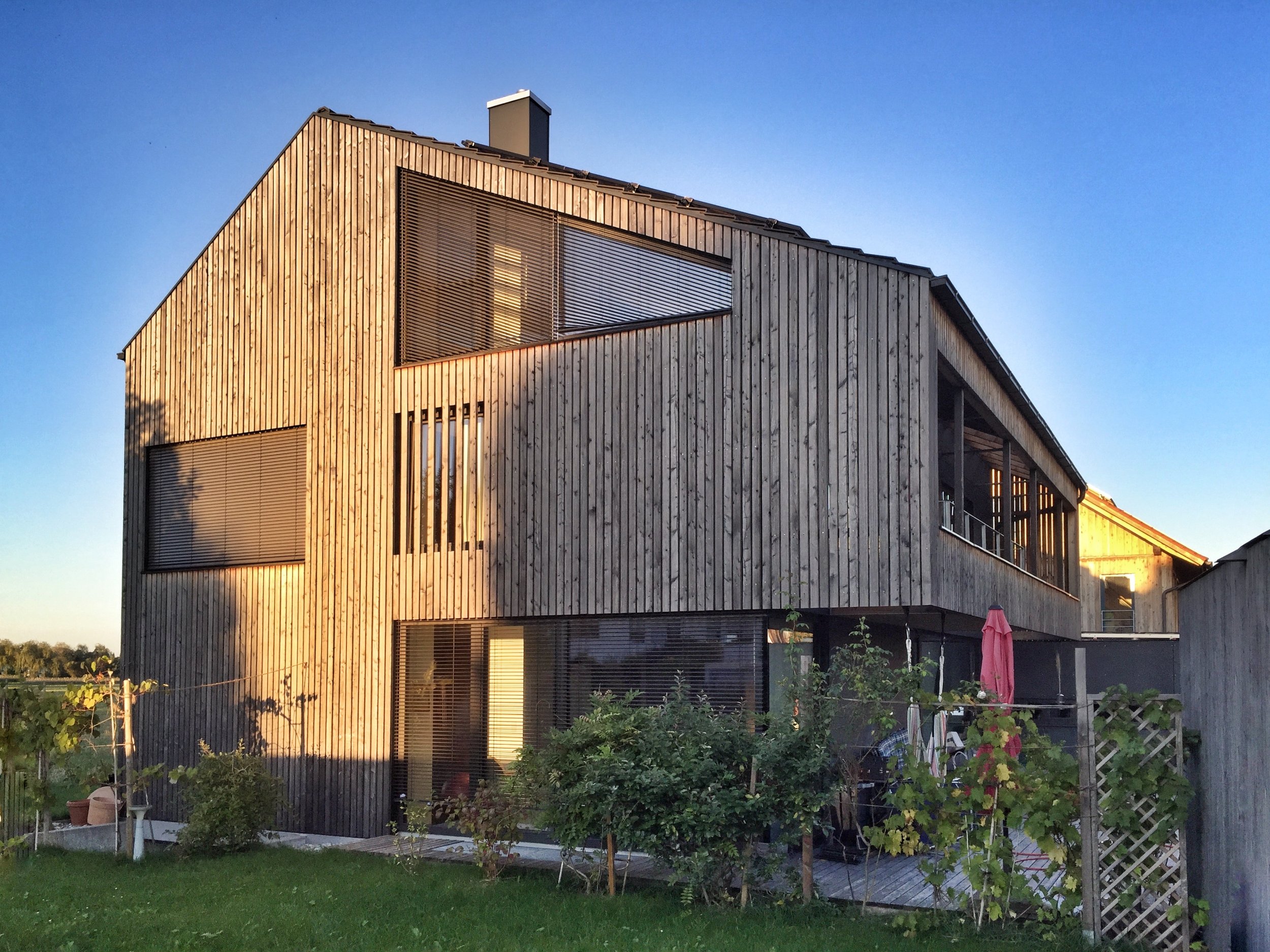 Modernes Mehrfamilienhaus mit Holzfassade, große Fenster und Balkon, im Garten sind Pflanzen und eine Terrasse zu sehen, unterklappbare Markise und Gartenmöbel