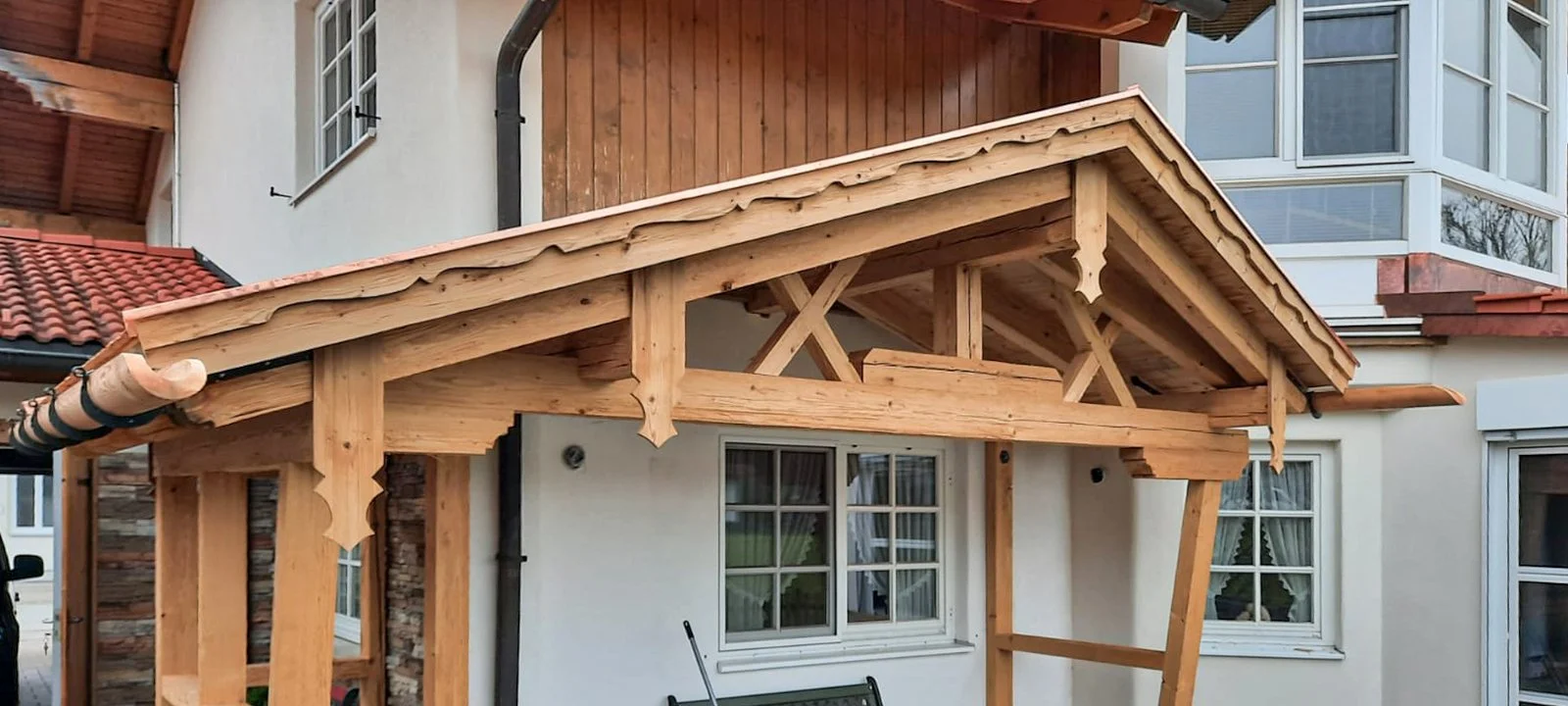 Holzüberdachung vor einem weißen Haus mit mehreren Fenstern und einem Balkon mit Balkonbrüstung.
