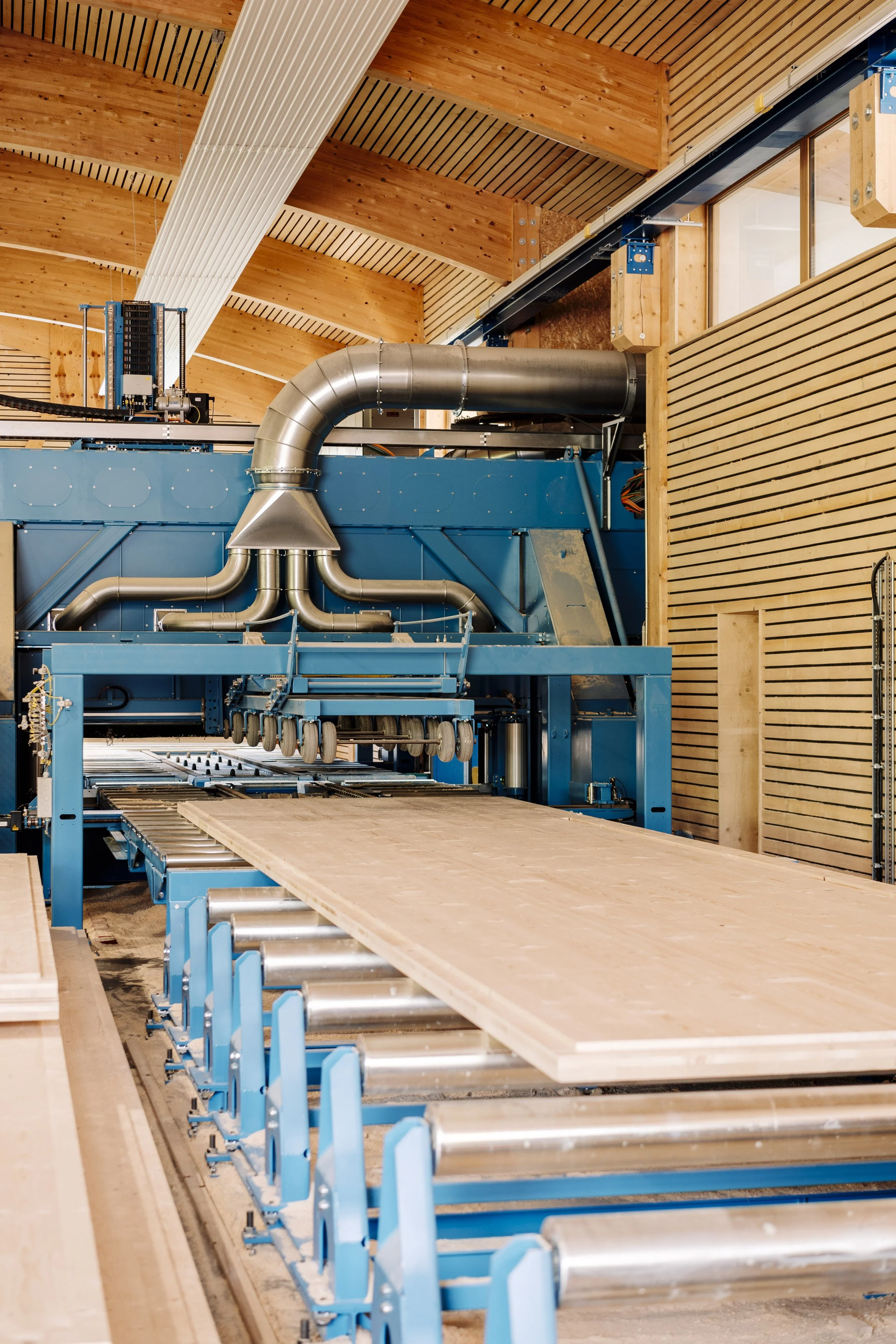 Ein industrieller Holzverarbeitungsraum mit einer großen blauen Maschine, die Holzplatten verarbeitet, und einem abgehängten Holzdeckenraum mit Fenstern.