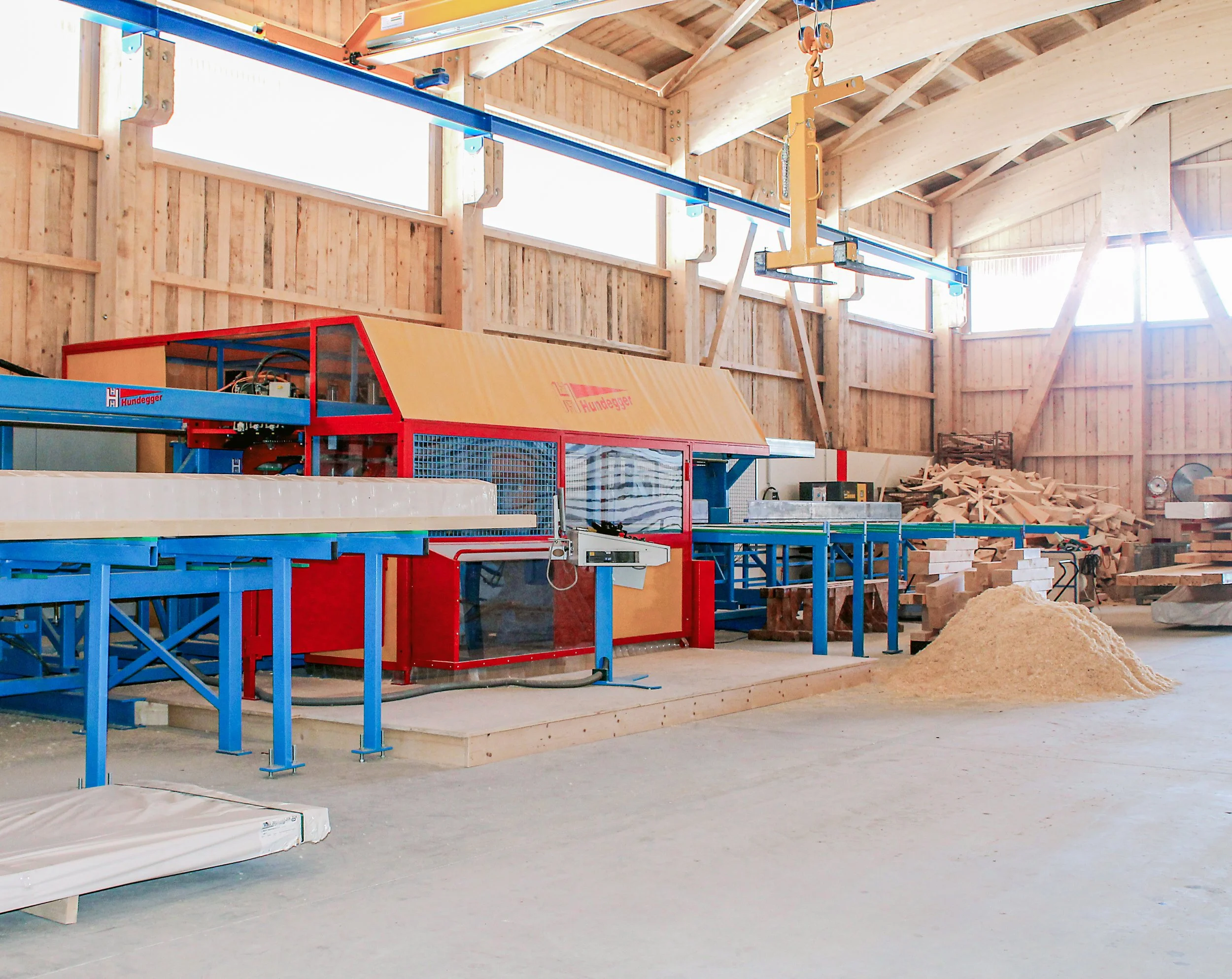 Innenraum einer Holzwerkstatt oder eines Sägewerks mit Holzbalken, Holzspänen, einer Maschine und Baugerüsten.