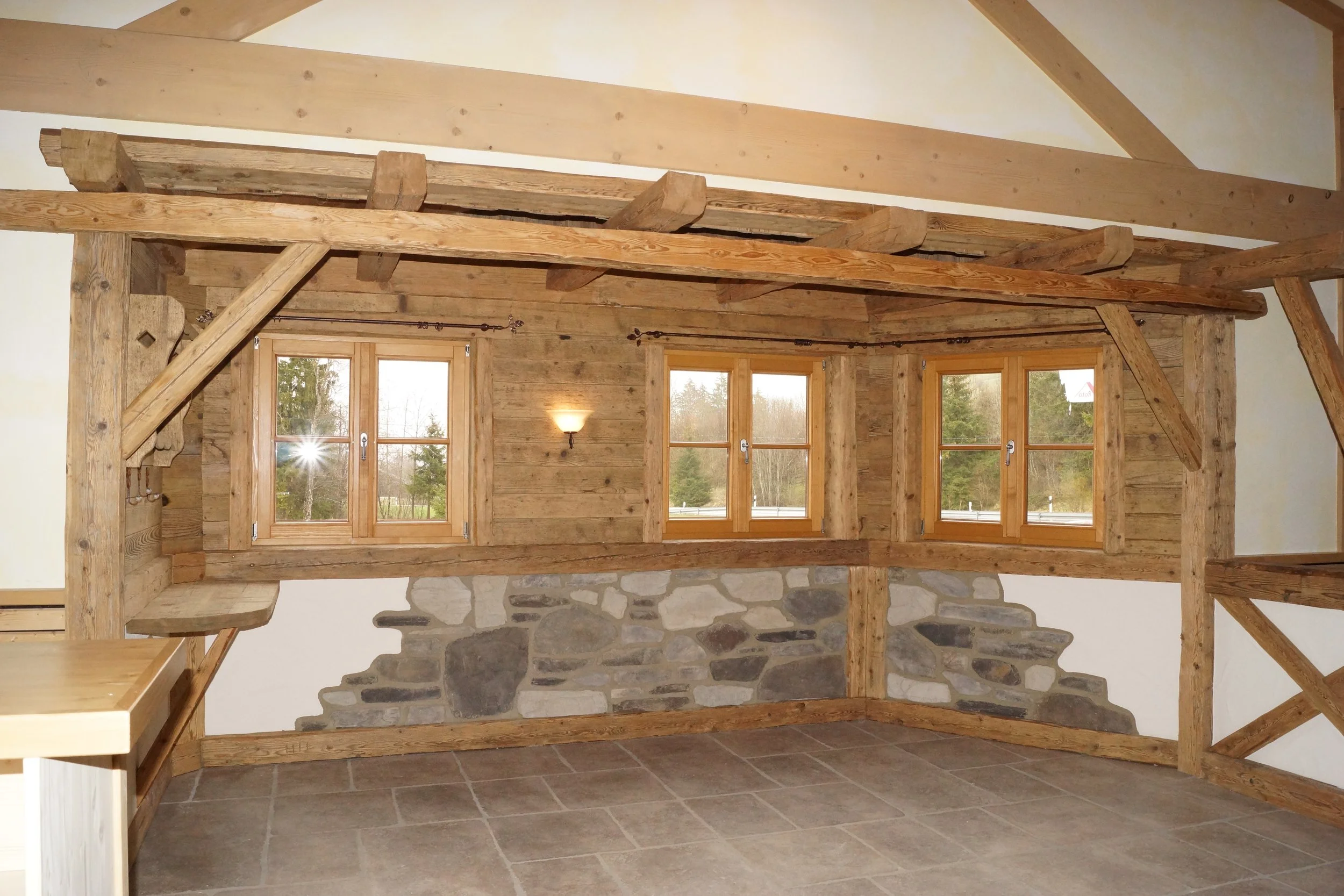 Innenaufnahme eines Raumes mit Holzwand und -decke, mehreren Fenstern, einem Steinwandabschnitt und einem Wandlicht