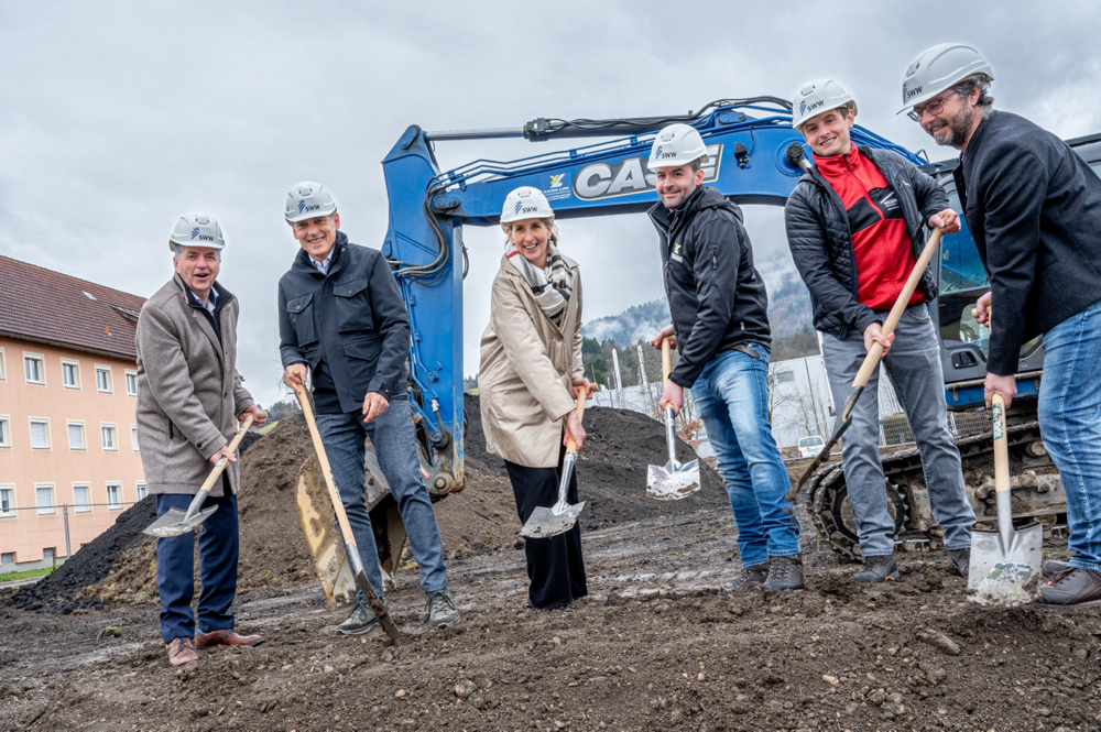 Fünf Menschen mit Bauhelmen beim Spatenstich auf einer Baustelle, inklusive Baufahrzeug im Hintergrund.
