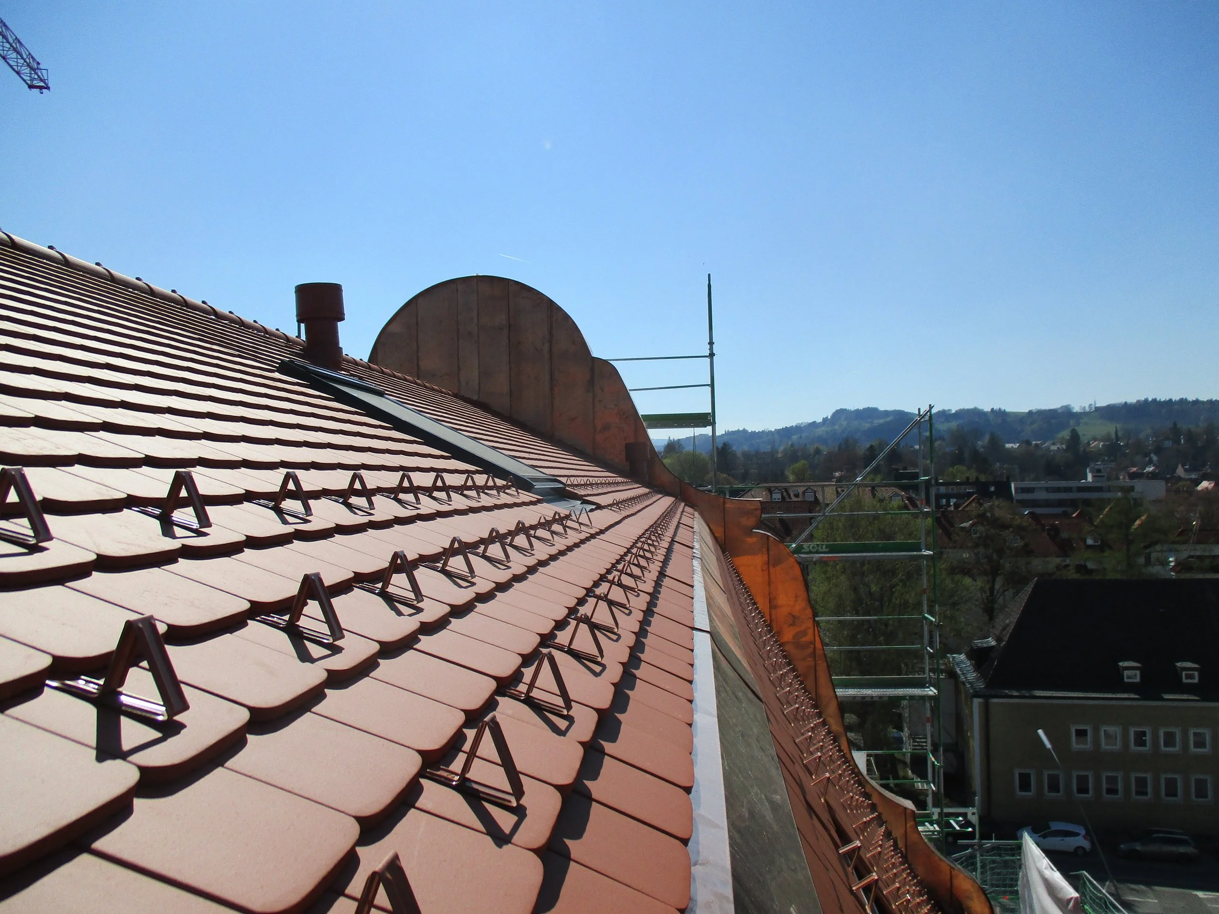 Dach mit roten Ziegeln, auf dem Baugerüst steht ein halbkreisförmiges Holzstück, im Hintergrund sind Berge und Häuser sichtbar, der Himmel ist klar und blau.