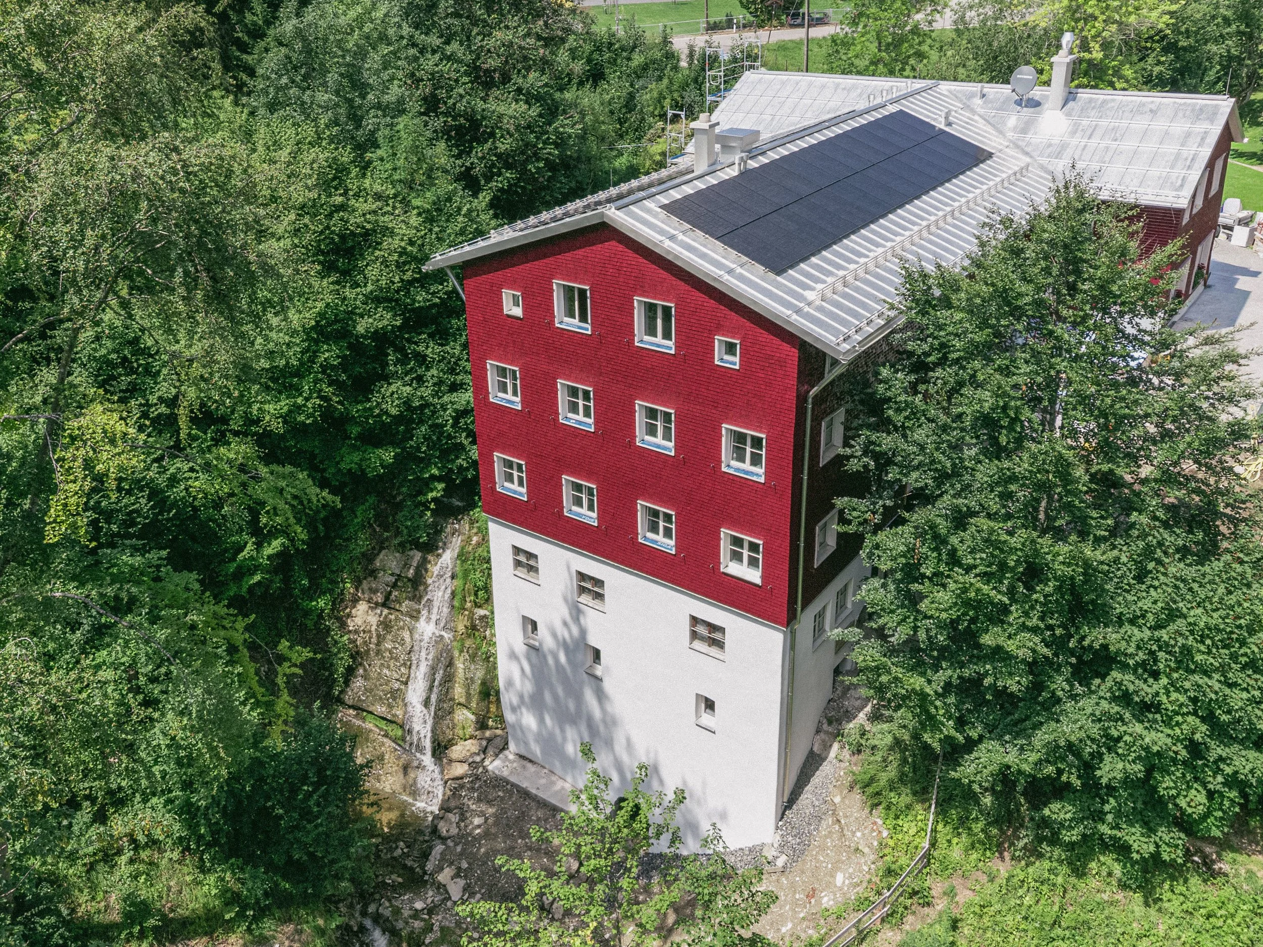 Ein Haus mit roten und weißen Wänden, das auf einem Felsen gebaut ist, ist von vielen grünen Bäumen umgeben. Das Dach hat Solarpaneele.