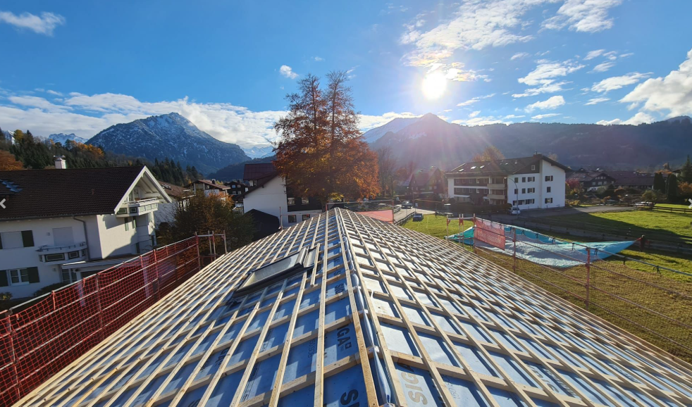 Blick auf ein Gebäude mit laufender Dachkonstruktion, im Hintergrund Berge, sonniger Himmel, Bäume mit Herbstlaub, und andere Gebäude in einer ländlichen Gegend.