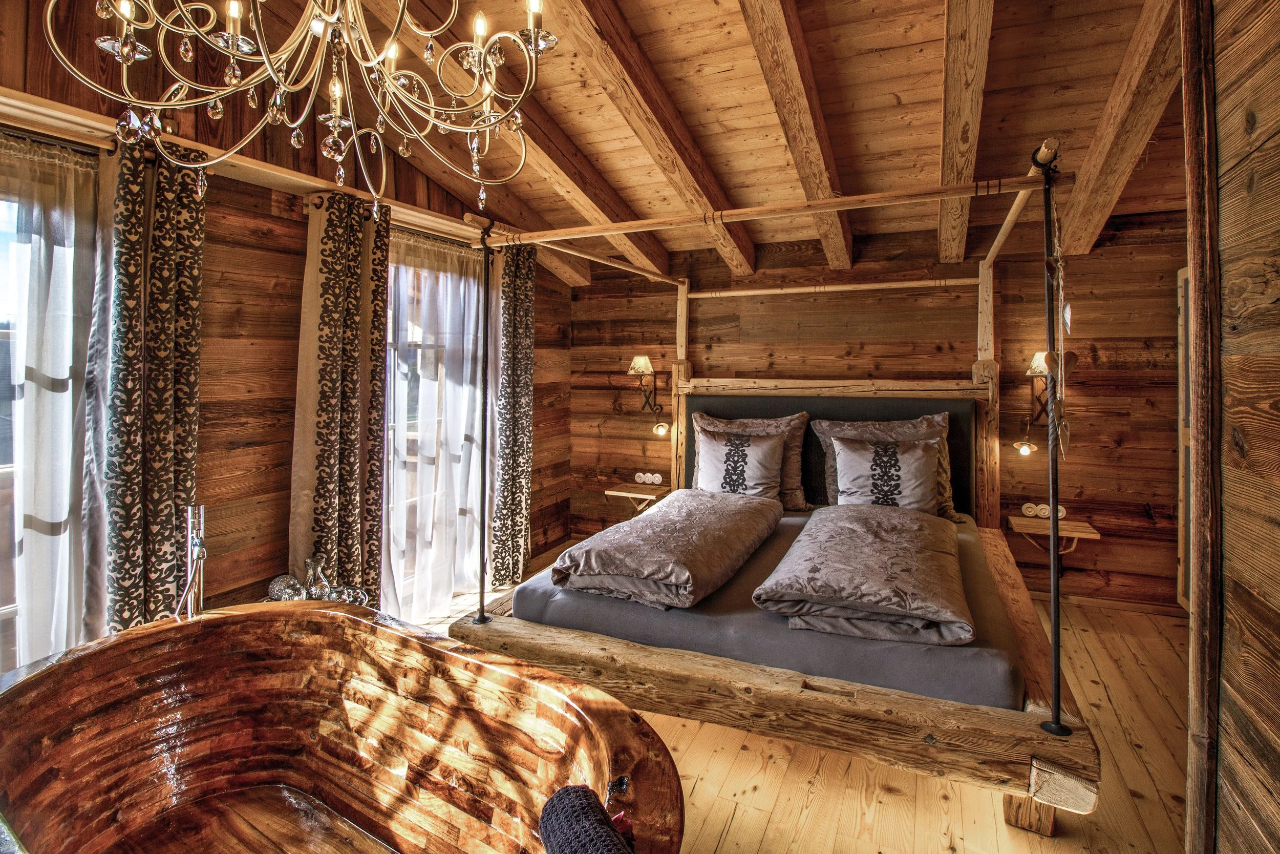 Ein Schlafzimmer mit Holzdecke und Holzverkleidung an den Wänden. Es gibt ein Doppelbett mit luftigen, silbergrauen Bettwäsche und Kissen, sowie eine Deckenlampe mit Kristallen. Die Fenster sind mit weißen und schwarzen Vorhängen geschmückt. Im Vorde