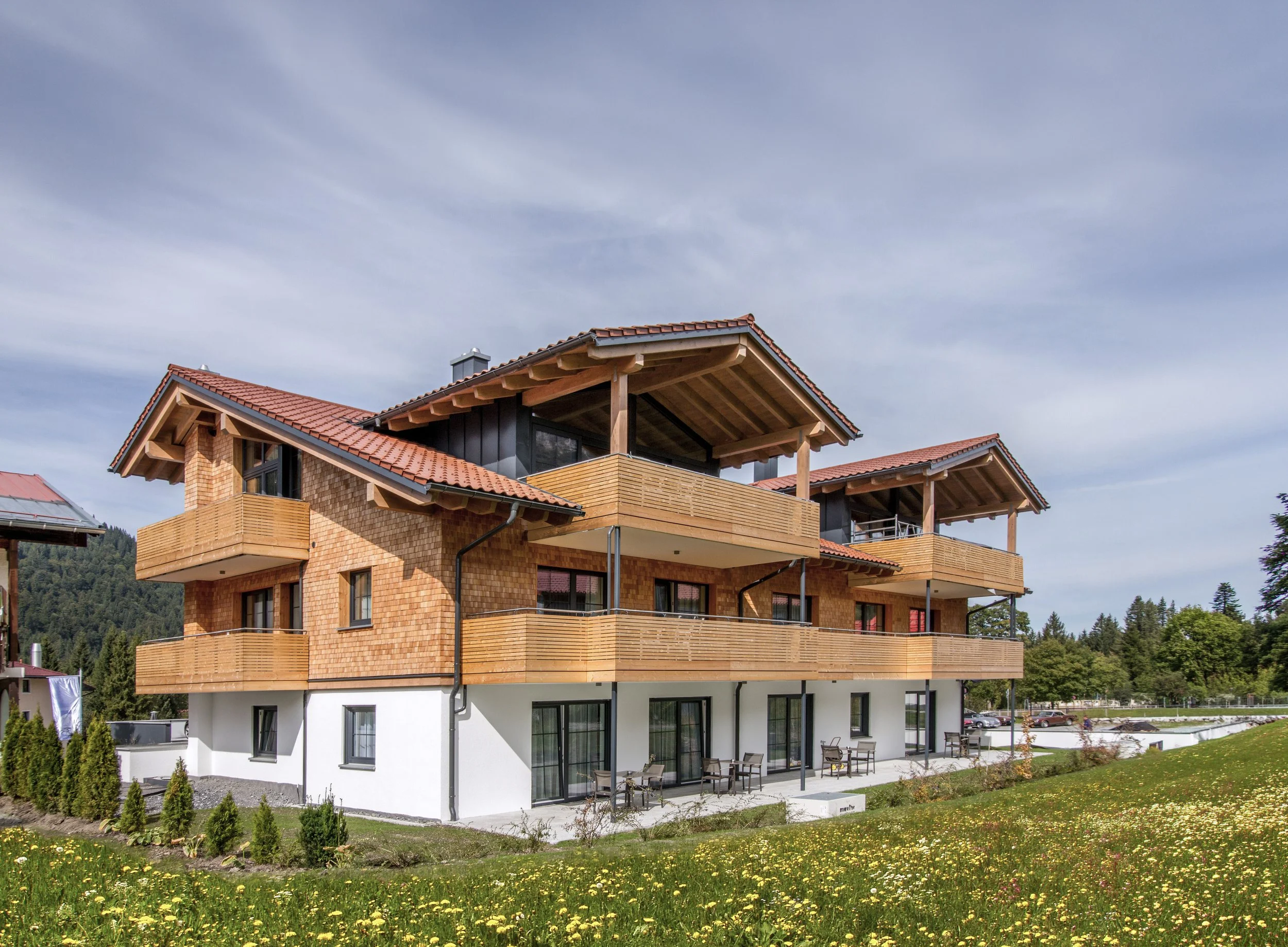 Ein modernes mehrstöckiges Haus mit terrakottafarbenen Ziegeldächern, Holzverkleidungen und großen Fenstern. Der Garten im Vordergrund ist grün mit Blumen und kleinen Pflanzen.