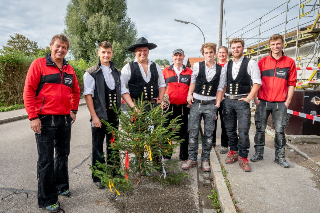 Menschen beim Weihnachtsbaumverkauf, einige in traditioneller Tracht, andere in sportlicher Kleidung, auf einer Straße bei bewölktem Himmel.