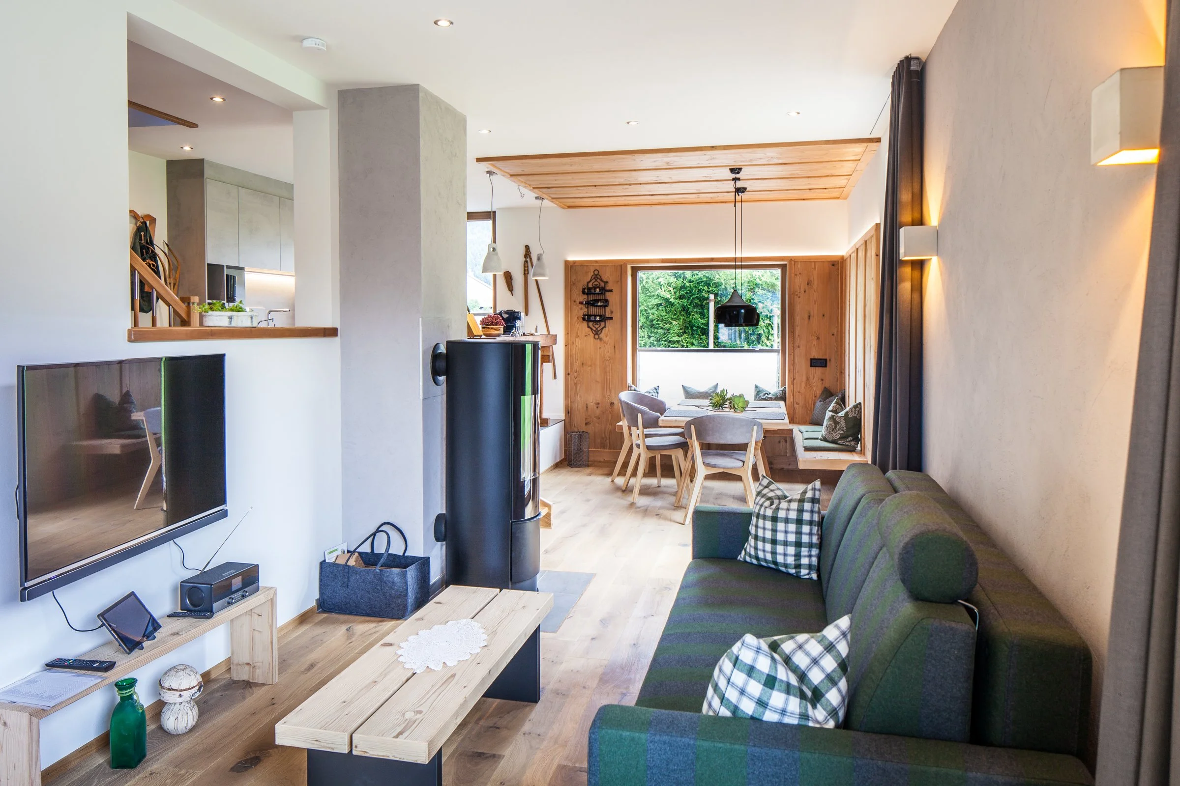 Offener Wohnbereich mit Esstisch, gemütlicher Couch, Holzverkleidung an der Wand, große Fenster mit Blick auf grünen Garten, moderne Beleuchtung, Fernseher auf Holzkonsole, Holzdecke im Essbereich.