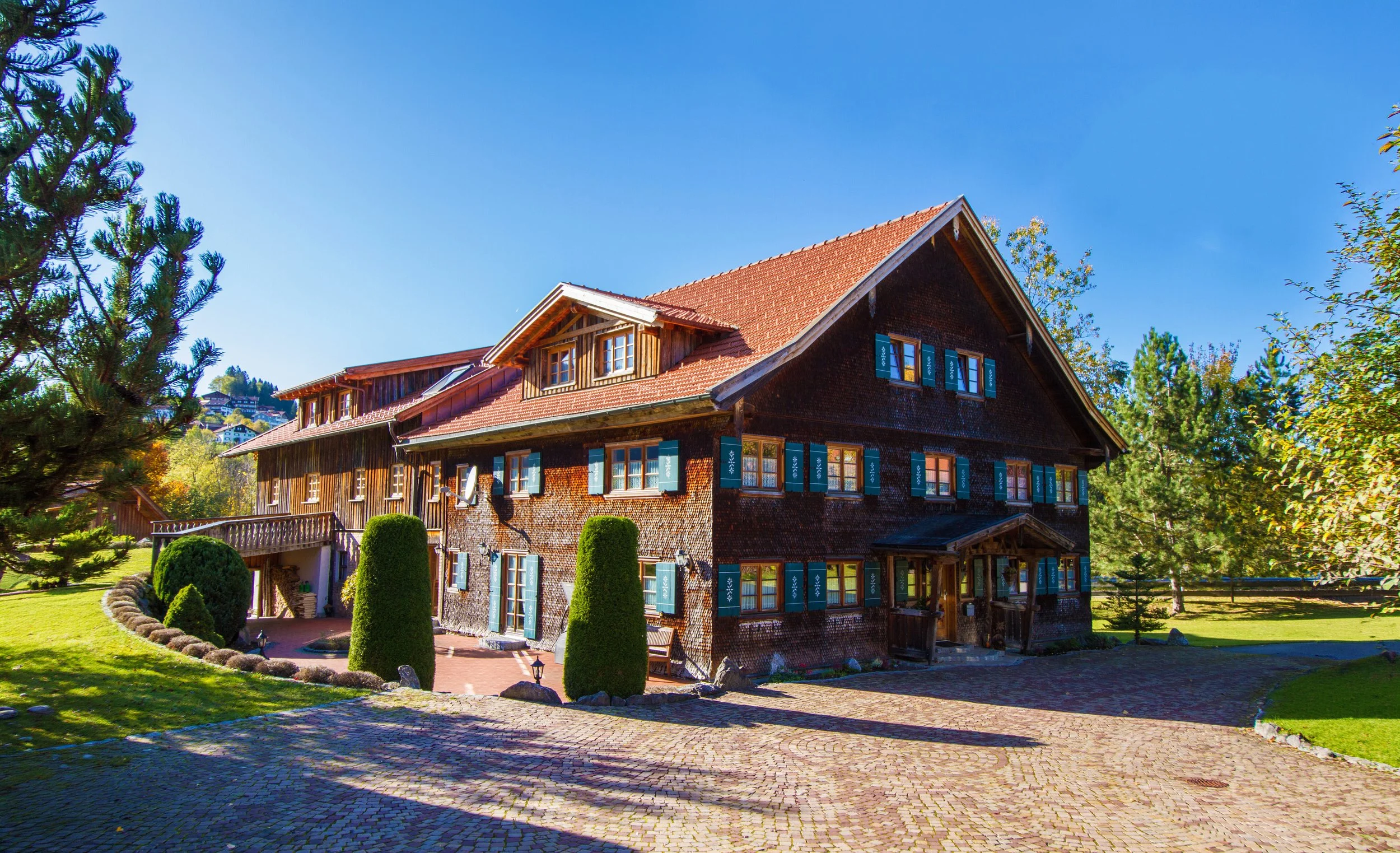 Ein großes, traditionelles Holzhaus mit roten Ziegel-Dach, grün gestrichenen Fensterläden, umgeben von einem gepflegten Garten mit Bäumen und Hecken, bei sonnigem Himmel.