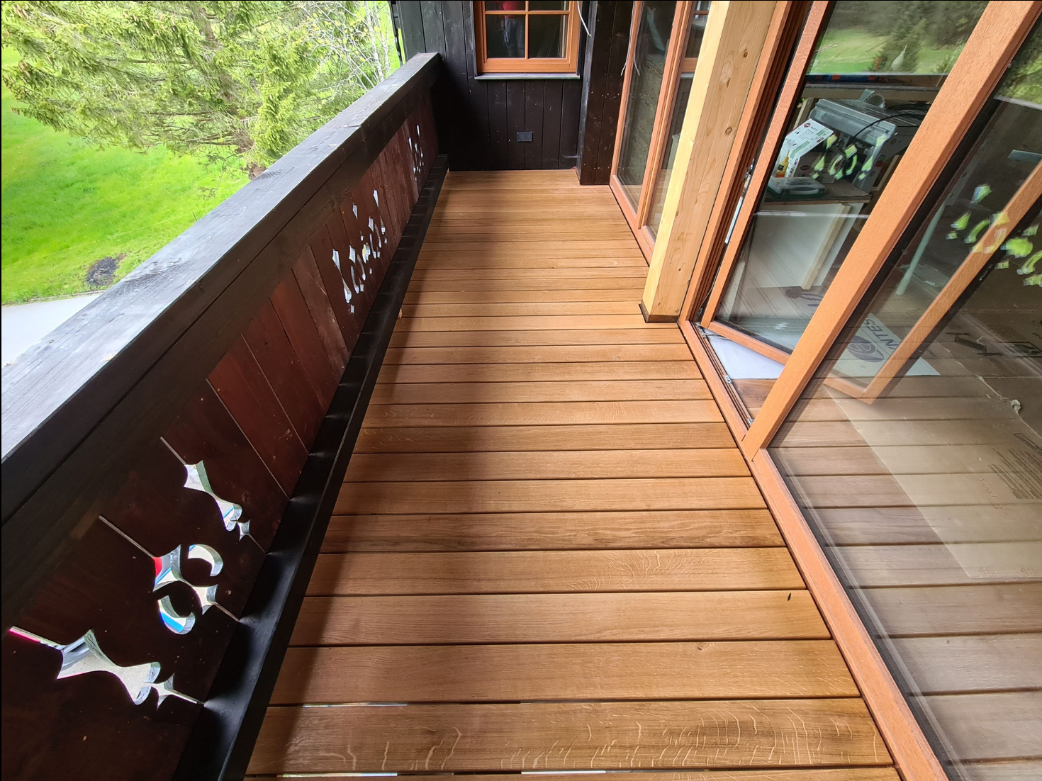 Balkon mit Holzfußboden und verzierten Holzgeländern, angrenzende große Glastüren führen ins Innere des Hauses, grüne Bäume im Hintergrund.