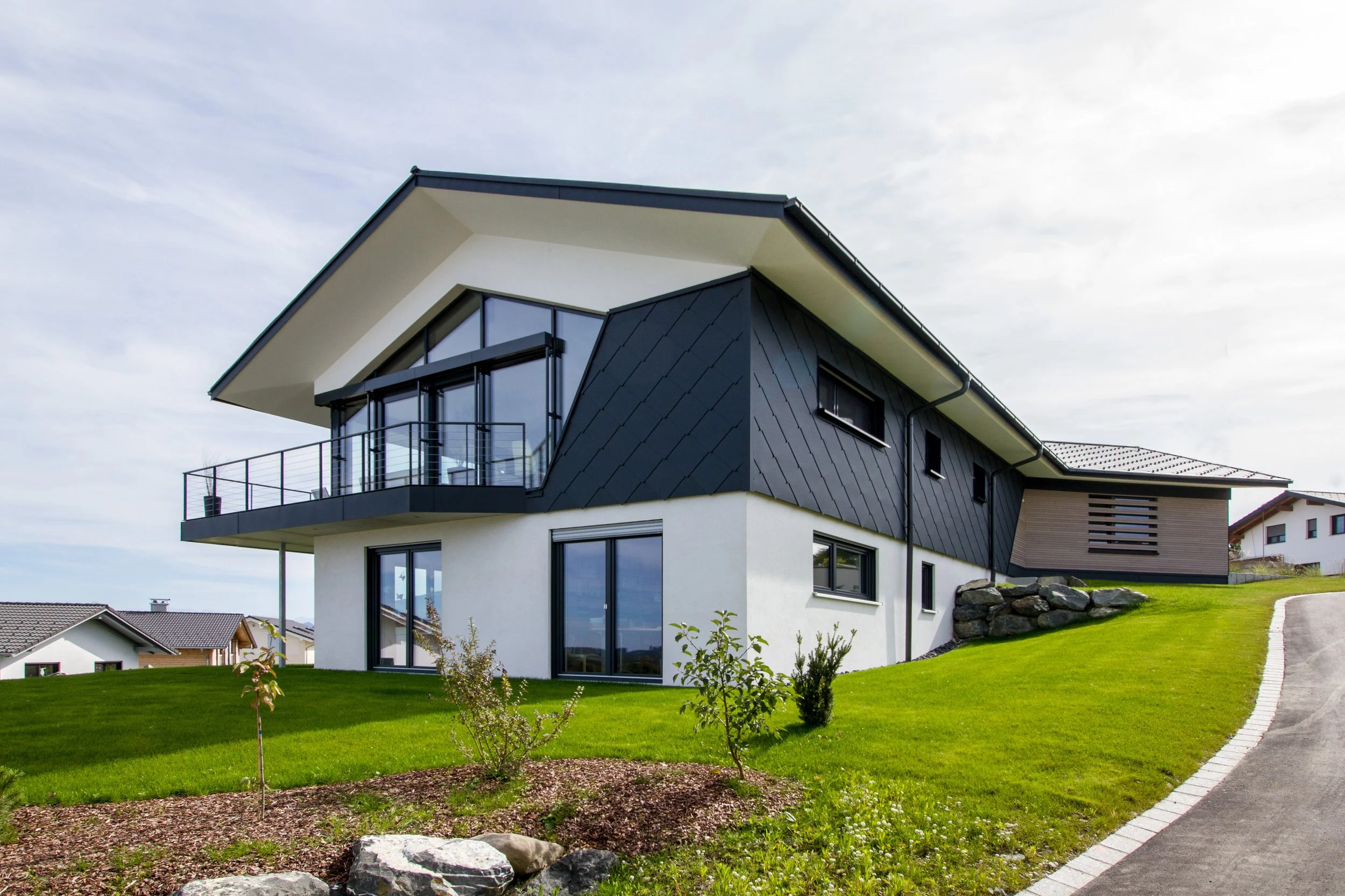 Modernes zweistöckiges Haus mit schwarzem Satteldach, großen Fenstern und einem Balkon, umgeben von einem gepflegten grünen Rasen und einer gepflasterten Wege. Himmel mit leichter Bewölkung.