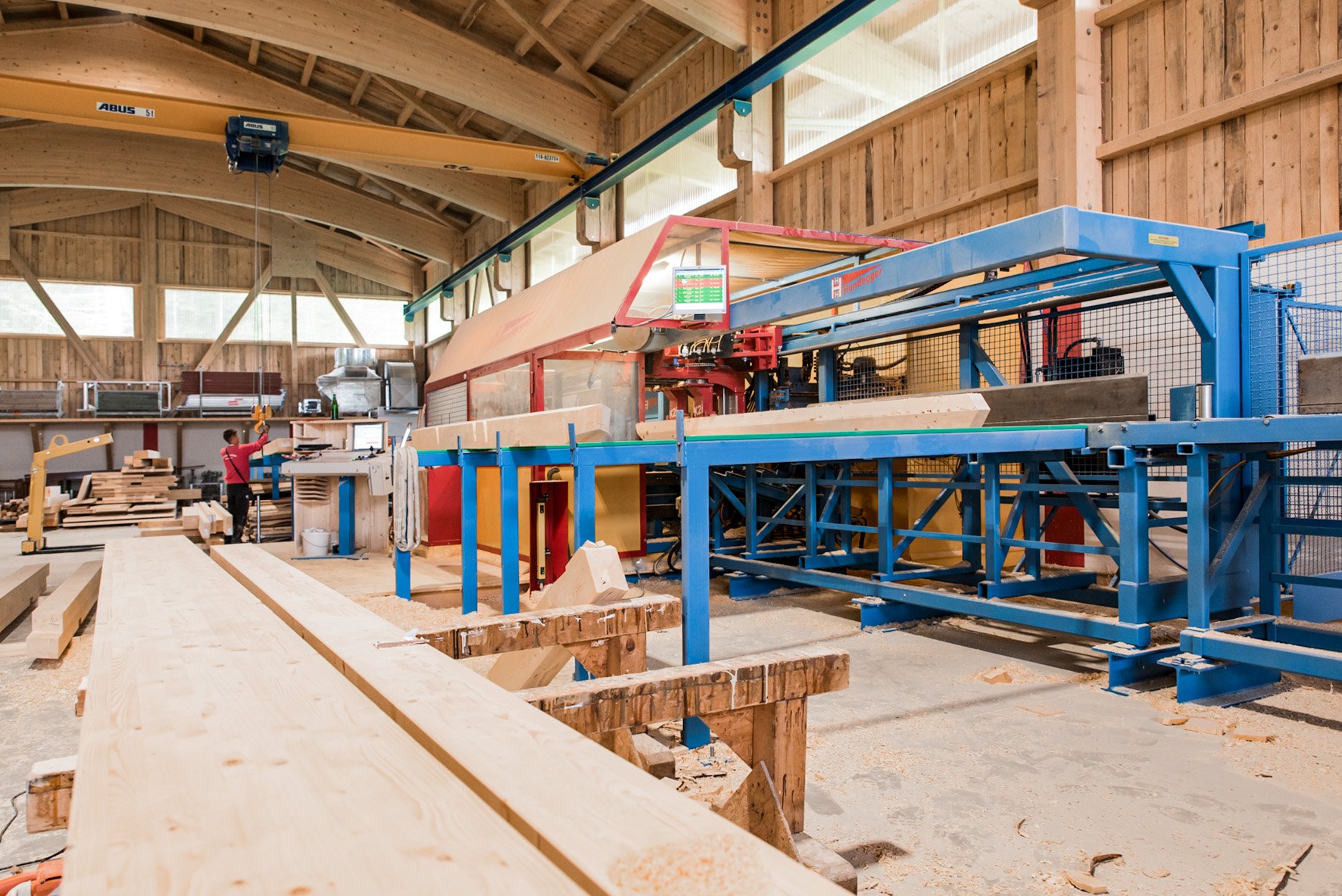 Holzverarbeitungsmaschine in einem Holzwerkstatt, mit einem Arbeiter, der Holzstücke verarbeitet, und Holzlatten im Vordergrund.