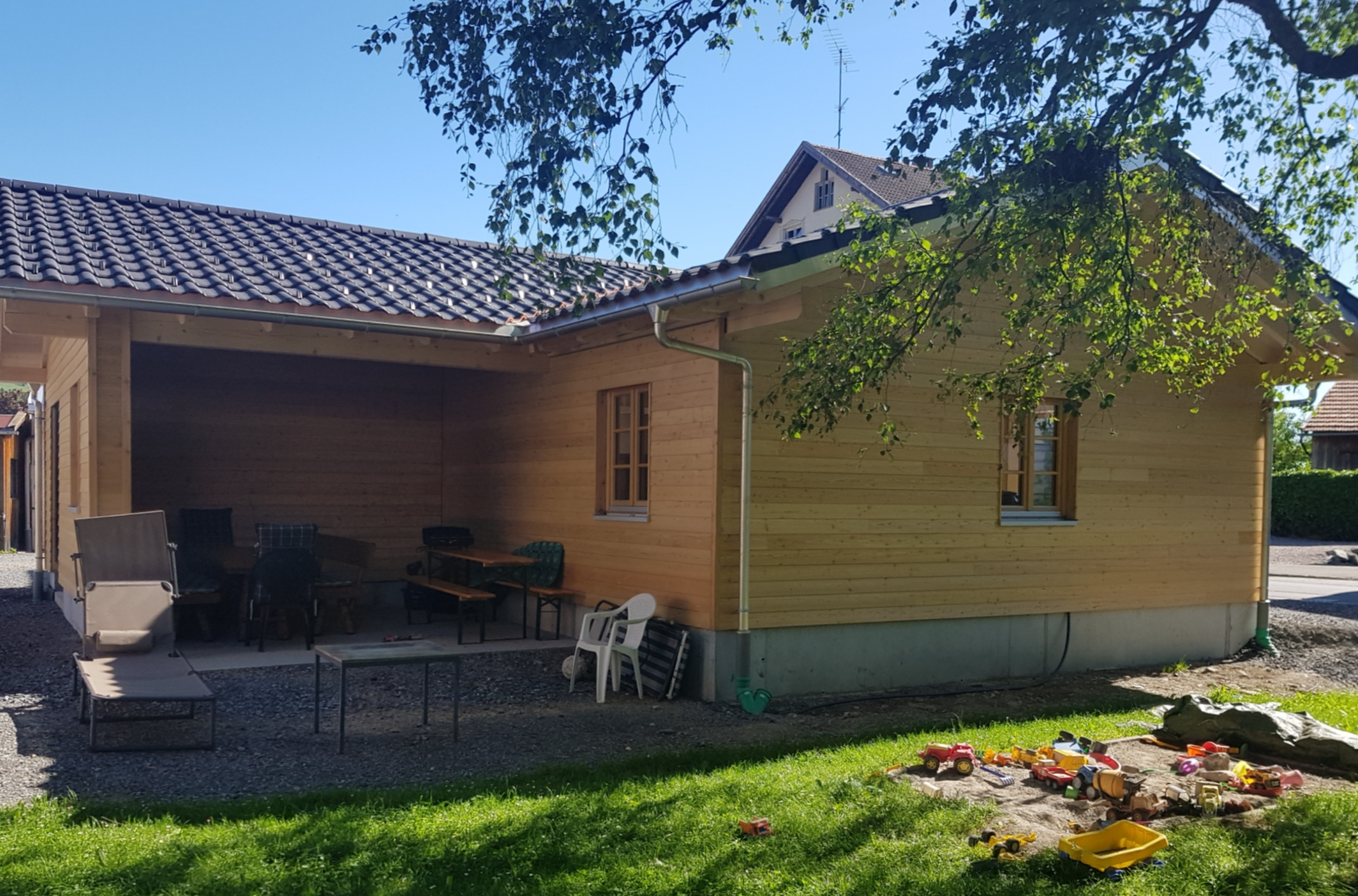 Ein Haus mit Holzwänden und roten Ziegeldach, im Vordergrund ein Garten mit Spielzeug und Möbeln, bei Sonnenschein, in den Bäumen blätter