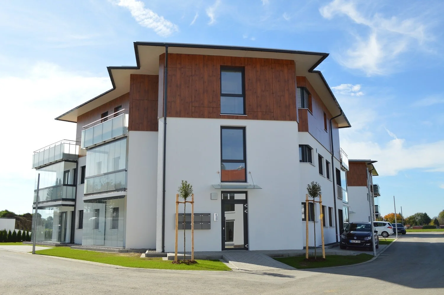 Moderne, mehrstöckige Wohnanlage mit weißen Fassaden, Holzverkleidung und Balkonen, im vorderen Bereich befinden sich kleine Bäume und Parkplätze.