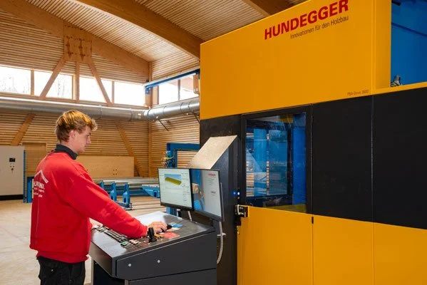 Ein junger Mann bedient eine Maschine in einer Werkshalle. Die Maschine ist gelb und schwarz und trägt den Namen 'HUNDEGGER'.
