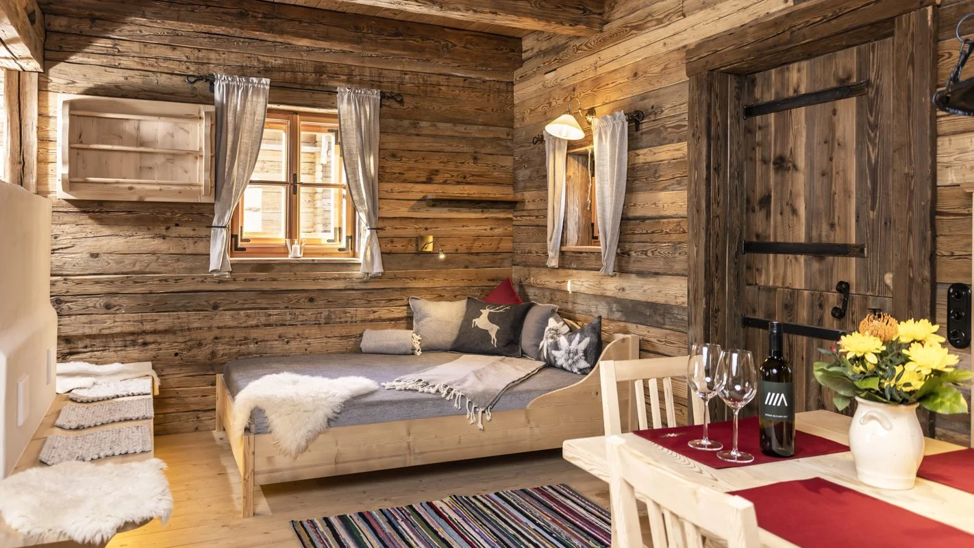 Gemütliches Holzhauszimmer mit Bett, zwei Fenster, Vorhänge, Tisch mit Wein und Blumen, rustikales Dekor.