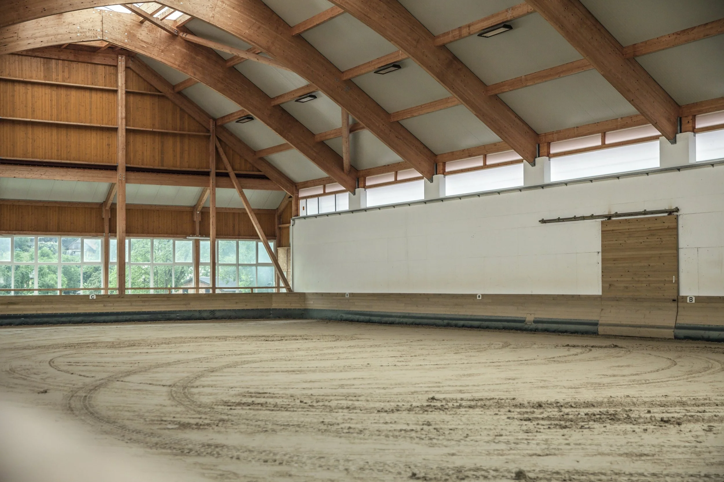 Innenraum einer großen Halle mit Holzbalkendecke, hohen Fenstern und einer darunterliegenden weißen Wand, die wahrscheinlich für eine Sporthalle oder eine ähnliche Nutzung gebaut wurde.